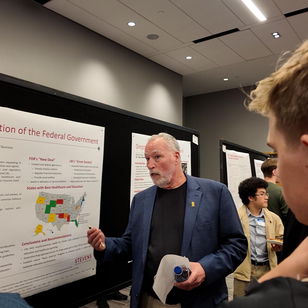 A professor in a navy blazer presents to students at a poster session, gesturing toward a research poster titled "Evolution of the Federal Government." Several students gather around listening, while other attendees view posters in the background.