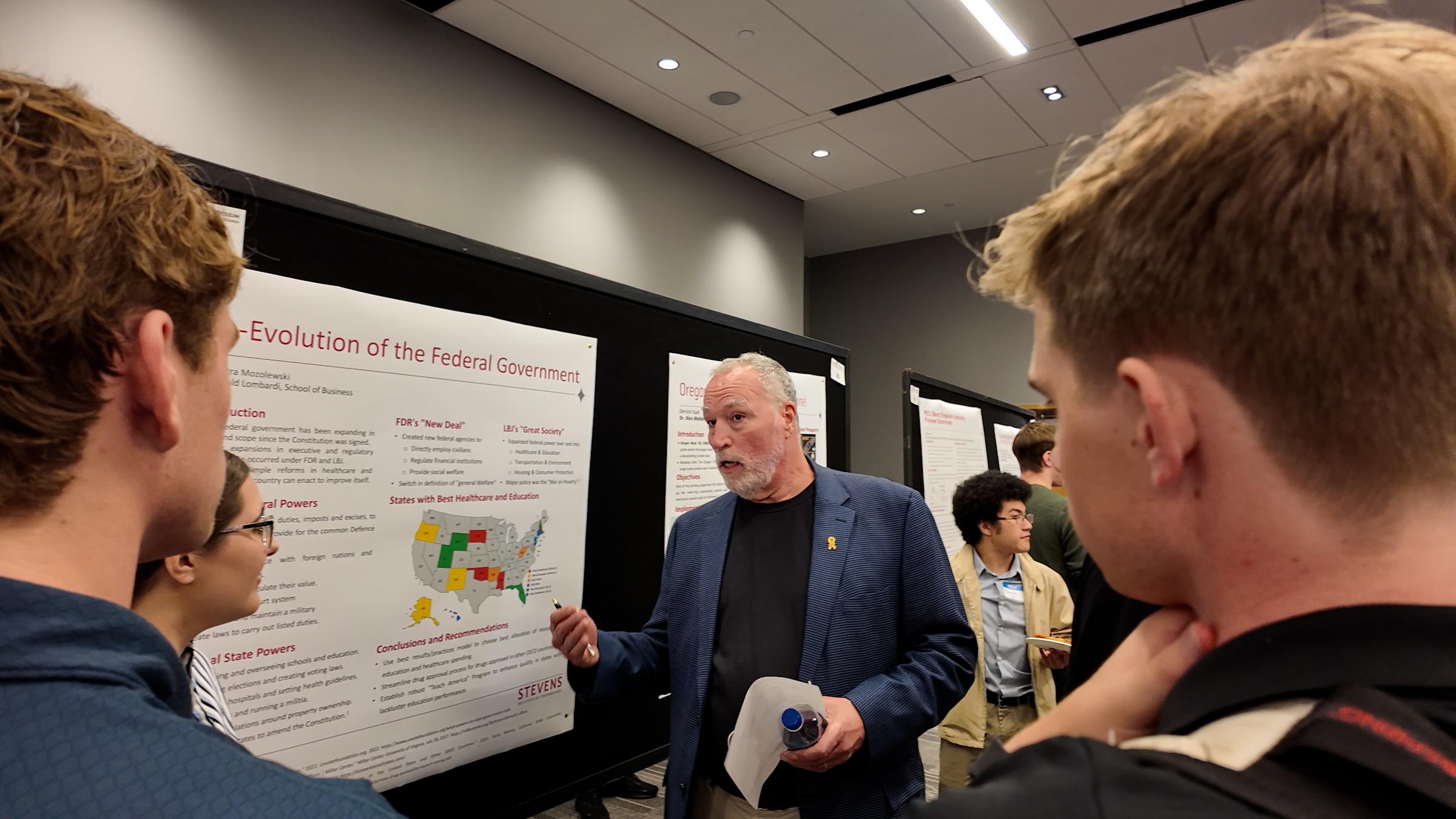 A professor in a navy blazer presents to students at a poster session, gesturing toward a research poster titled "Evolution of the Federal Government." Several students gather around listening, while other attendees view posters in the background.