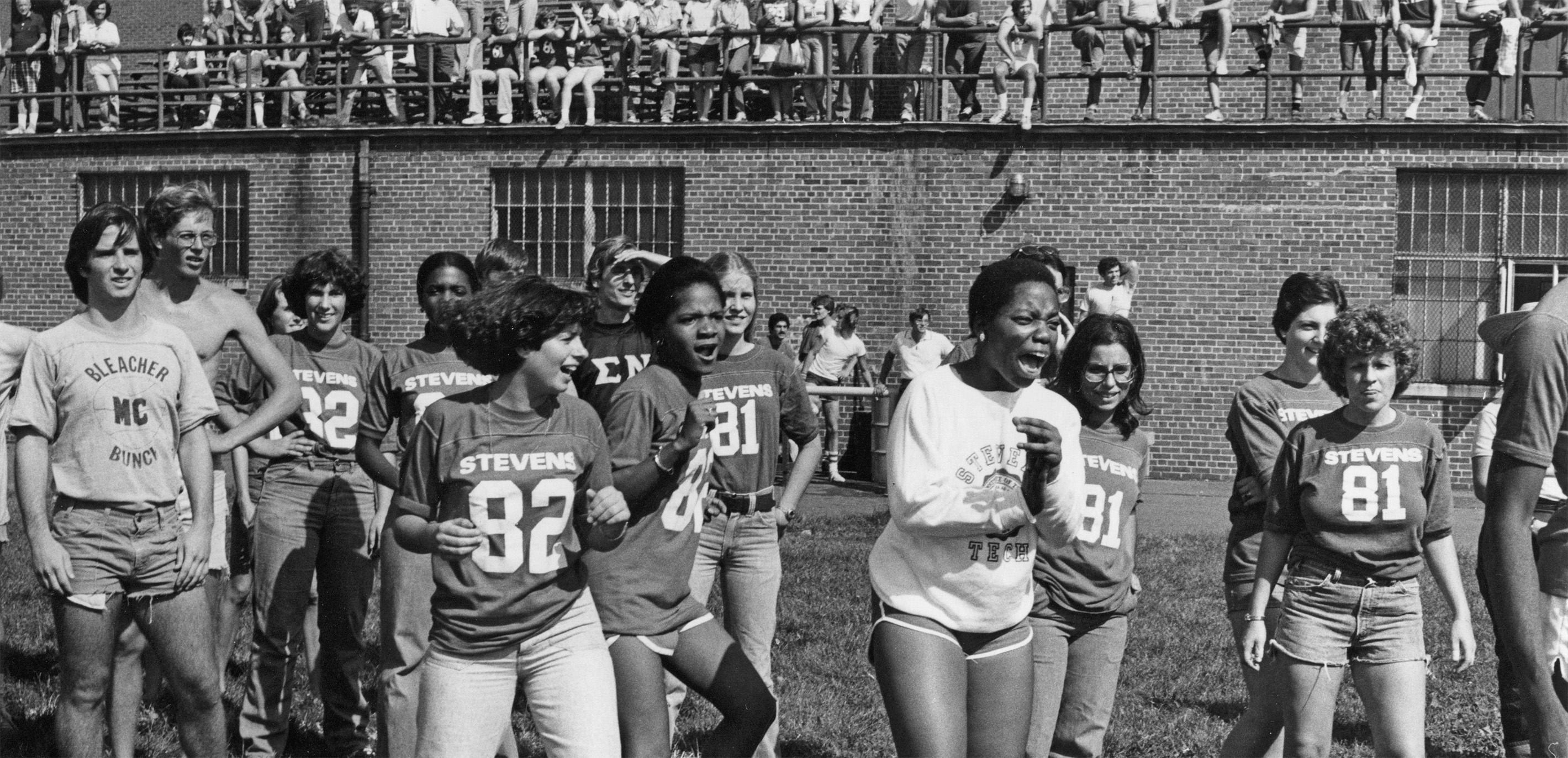 Large photo of a group of female students dancing and smiling on the field in front of the old Mott Field House, with groups of students watching from the bleachers.