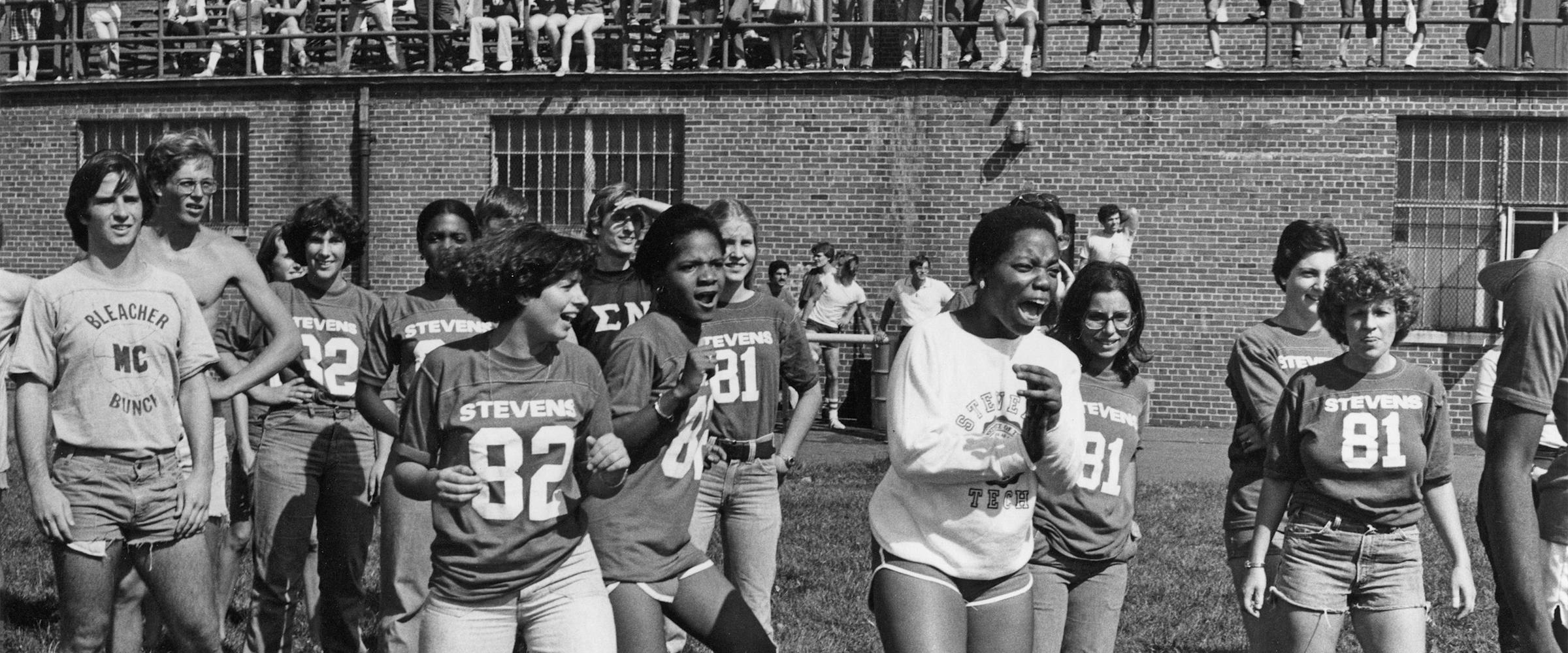 Large photo of a group of female students dancing and smiling on the field in front of the old Mott Field House, with groups of students watching from the bleachers.