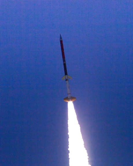 Nine Stevens students stand in front of a concrete partition. Behind them is a bridge. Suspended below the bridge is a rocket spanning the width of the photo.