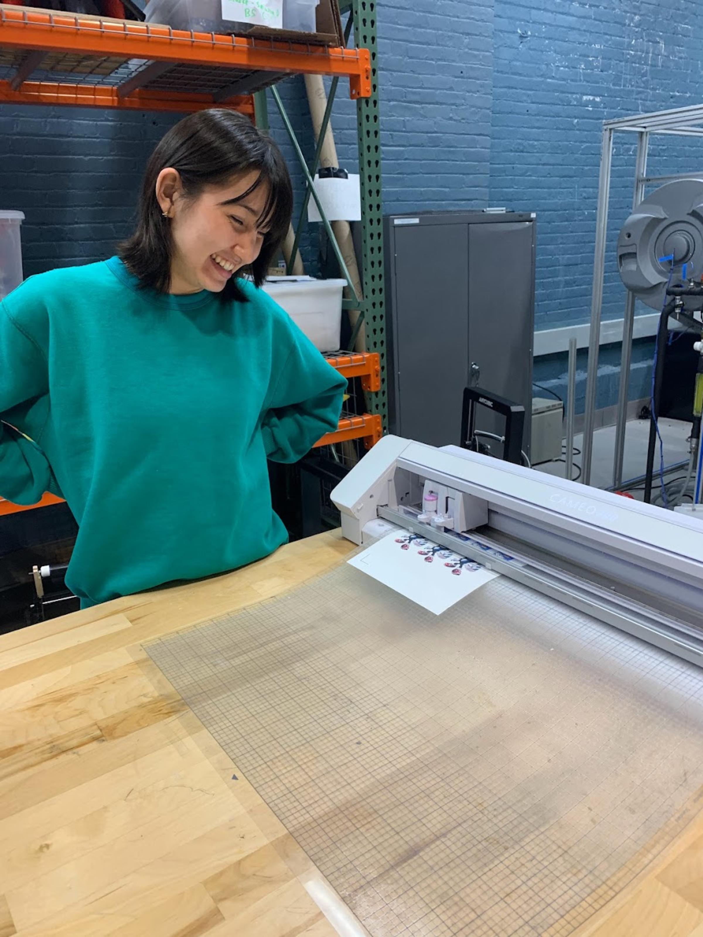 Female student wearing green shirt stands in front of a table that is printing out a plastic material in MakerSpace