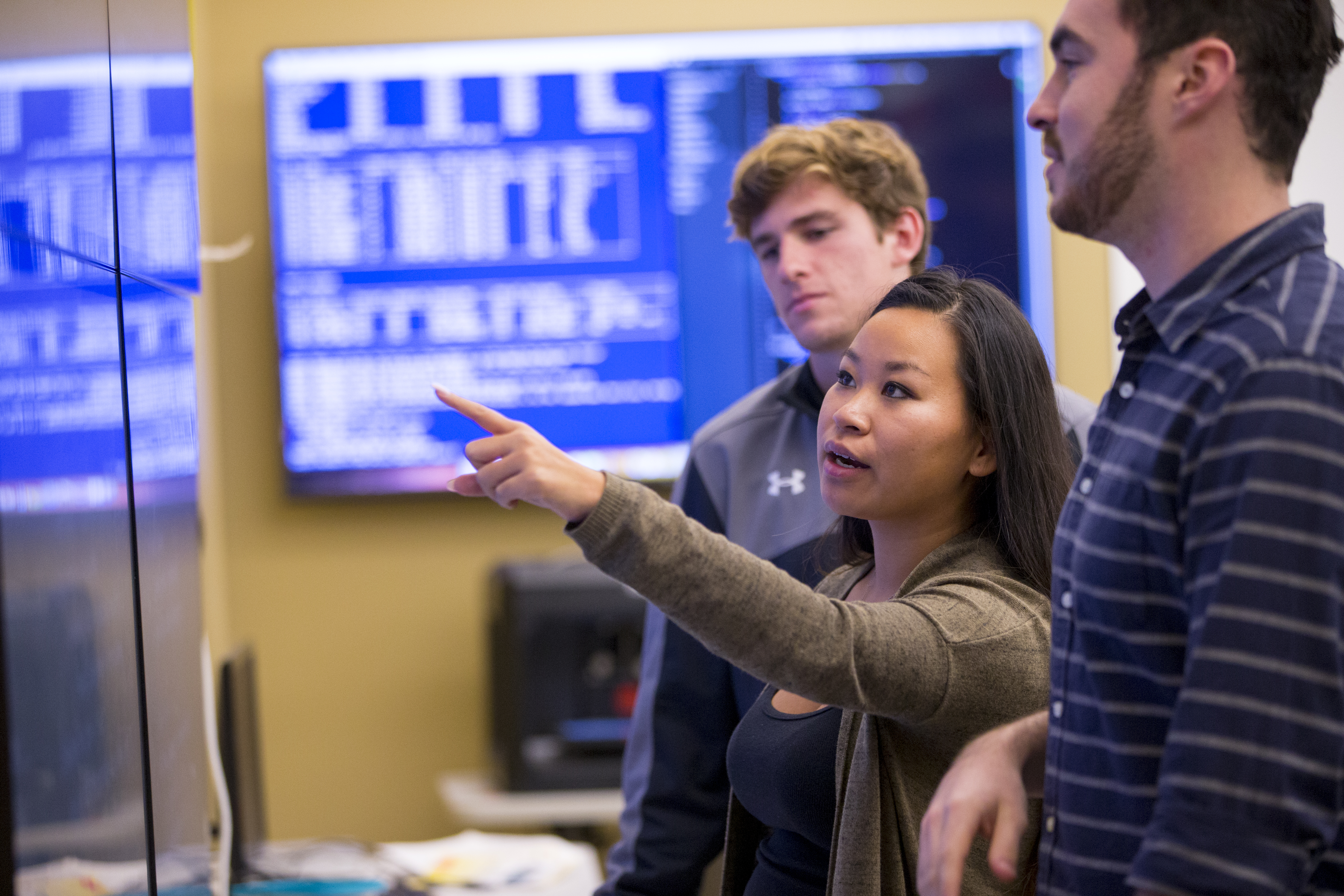 Student points at wall monitor while two other students look on.