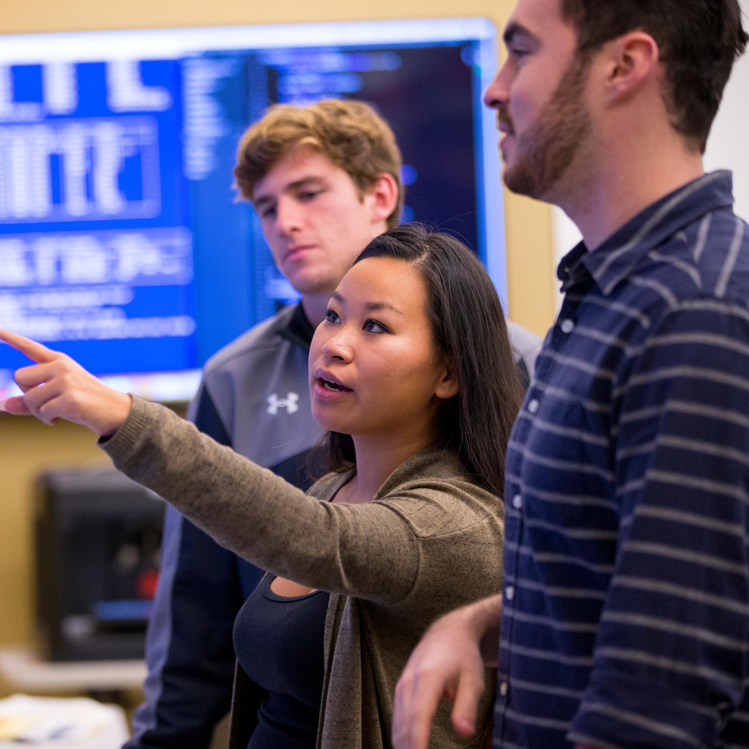 Student points at wall monitor while two other students look on.