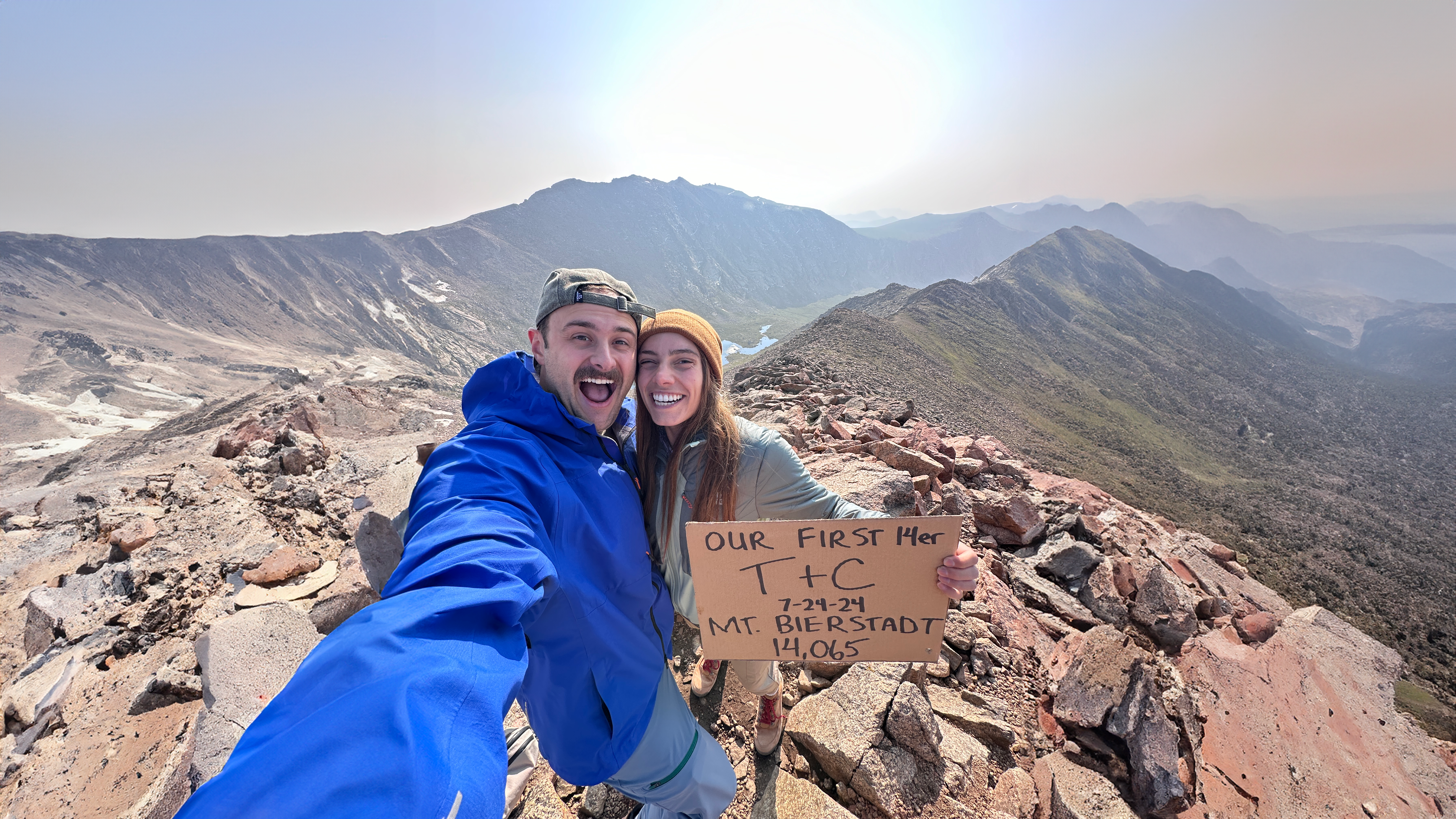 Two hikers celebrate at the summit of Mt. Bierstadt (14,065 ft) on July 24, 2024, holding a handmade sign reading "Our First 14er — T+C." They smile for a selfie with sweeping Rocky Mountain views behind them.