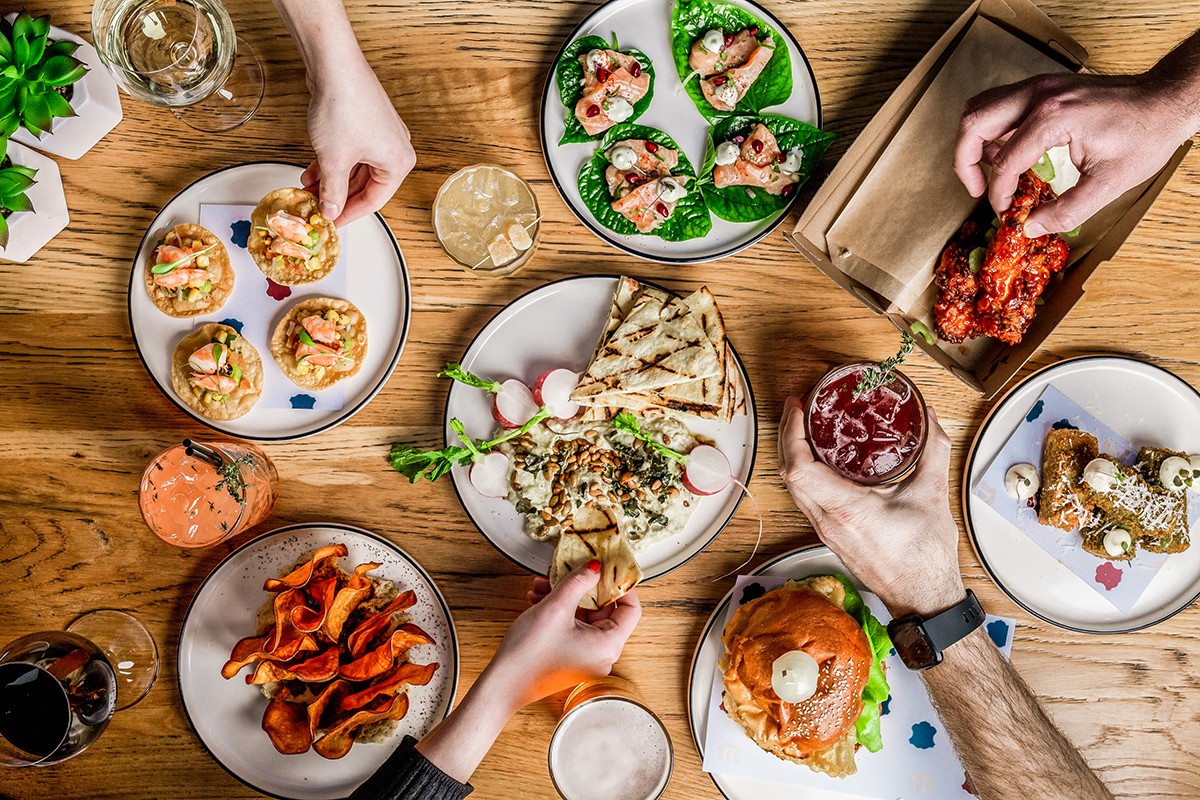 A wooden table with several white plates with food on them. Four people are touching the food with their hands.