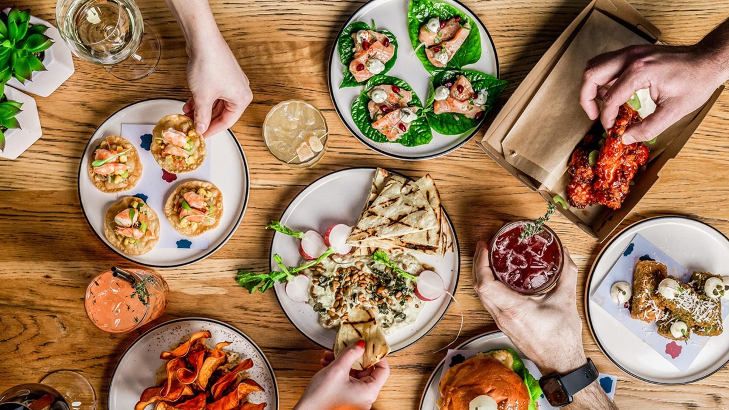 A wooden table with several white plates with food on them. Four people are touching the food with their hands.