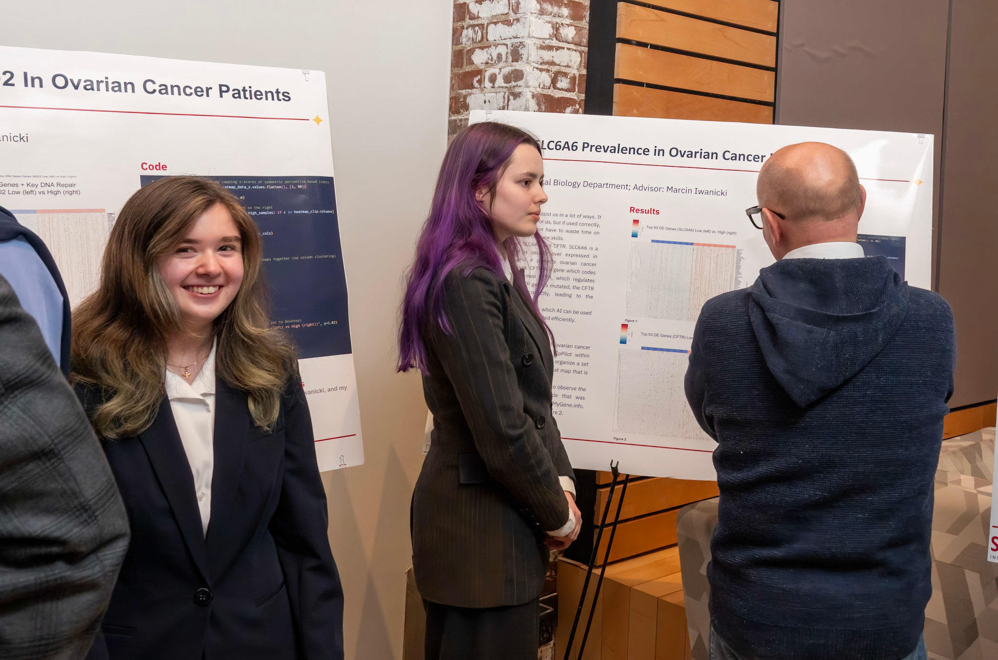 A smiling student at left and another student stand in front of presentation posters while attendees study them.