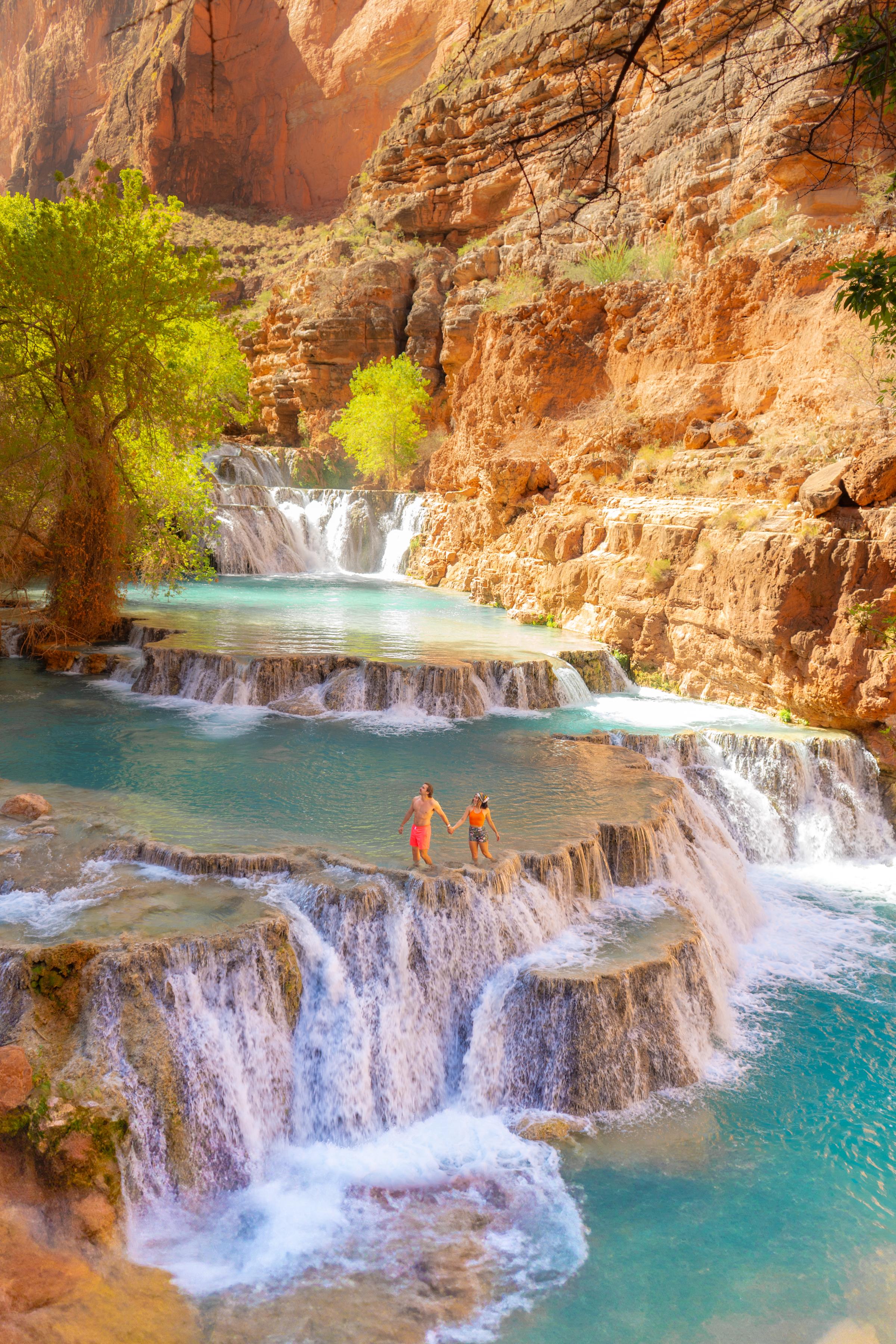 A couple holds hands while standing on tiered travertine waterfalls with vivid turquoise pools cascading down red sandstone canyon walls. Bright green trees contrast against the warm rust-colored rock in what appears to be Havasupai Falls, Arizona.