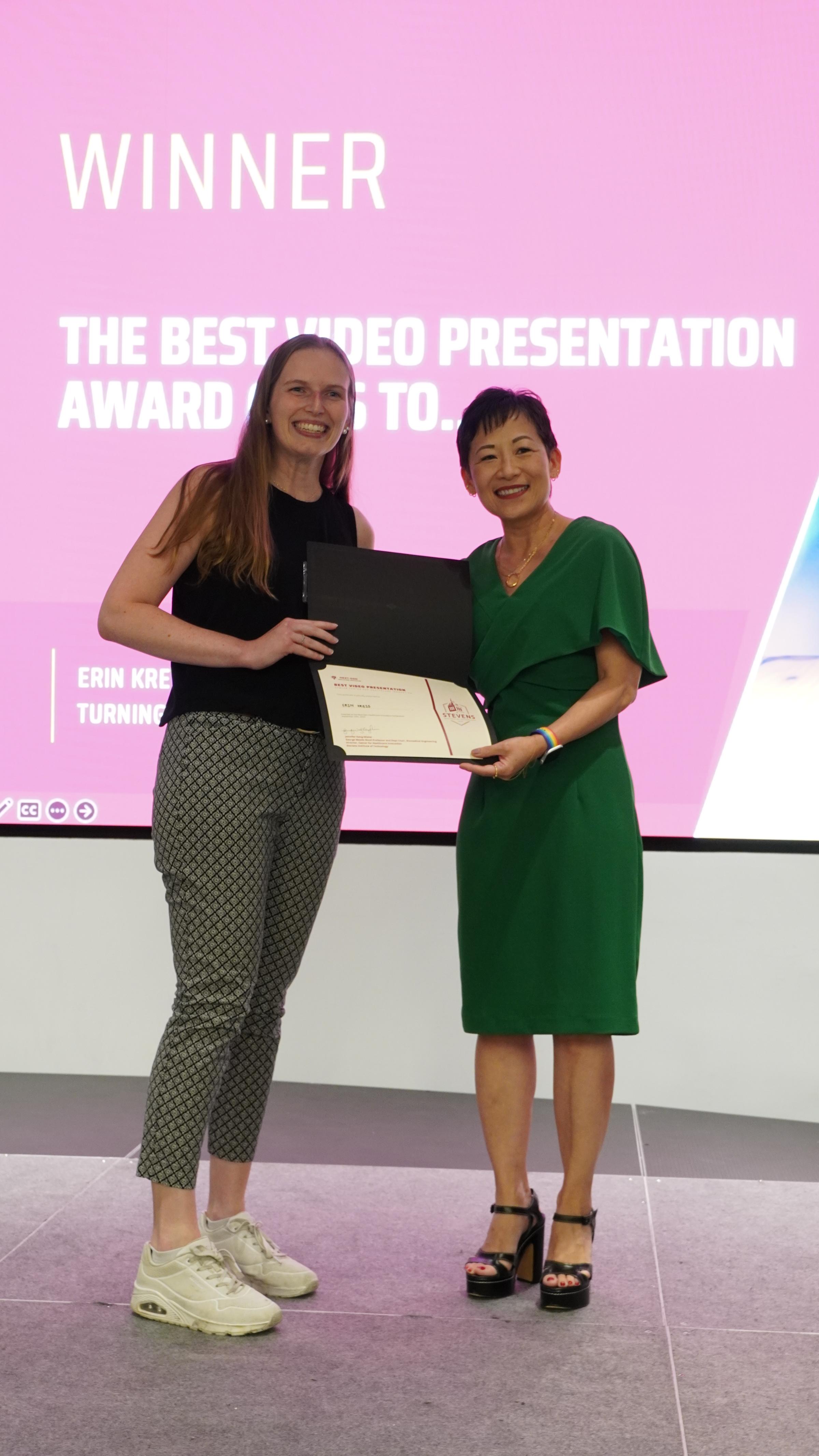 Erin Kries holds a commemorative winning certificate on a stage with Dr. Jennifer Kang-Mieler.