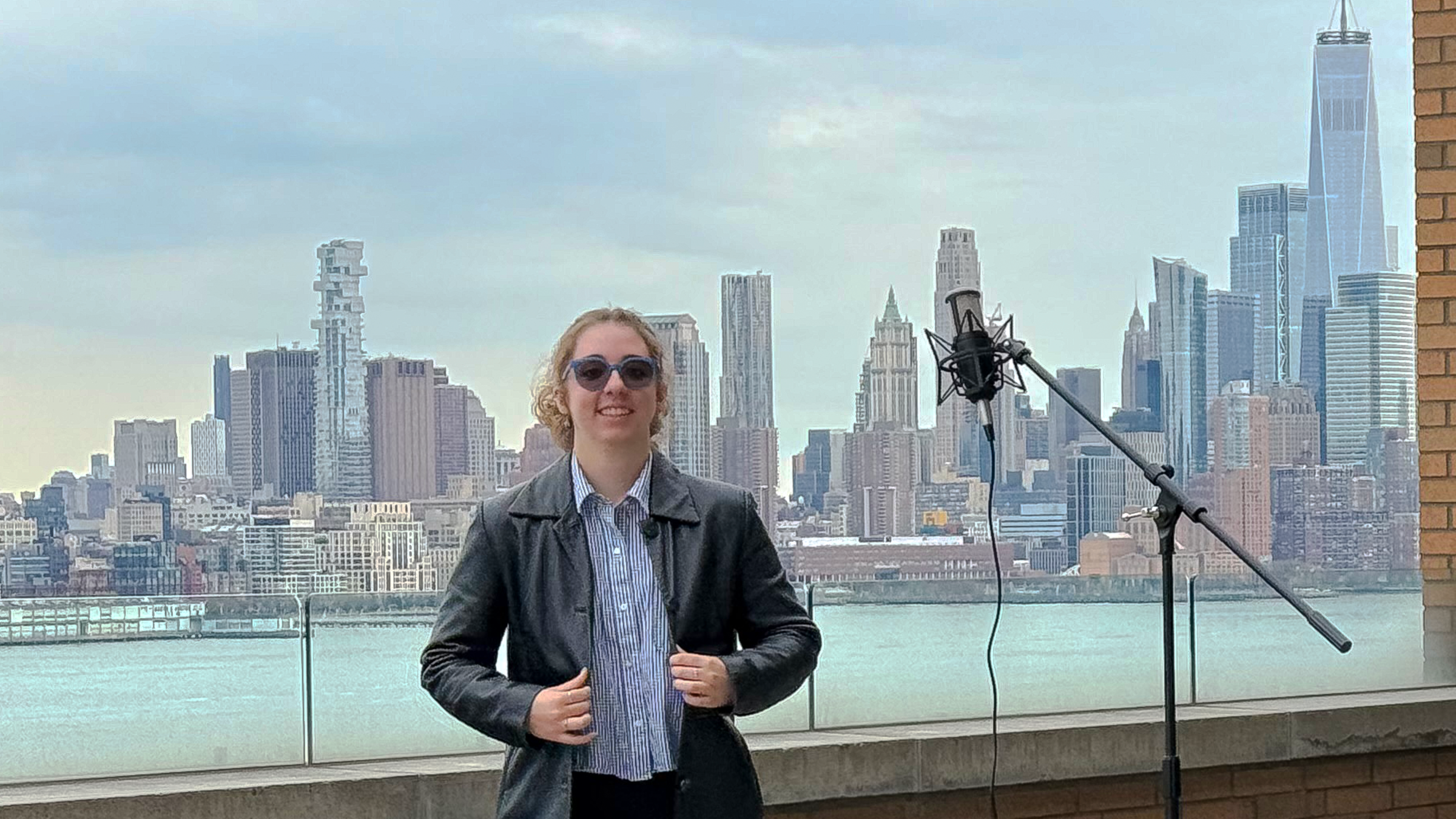A young woman with sunglasses and a leather jacket poses on Stevens' campus with the New York City skyline in the background.