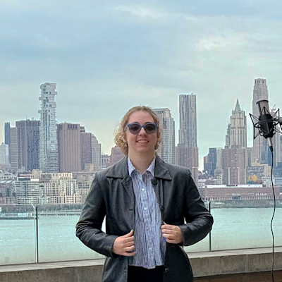 A young woman with sunglasses and a leather jacket poses on Stevens' campus with the New York City skyline in the background.