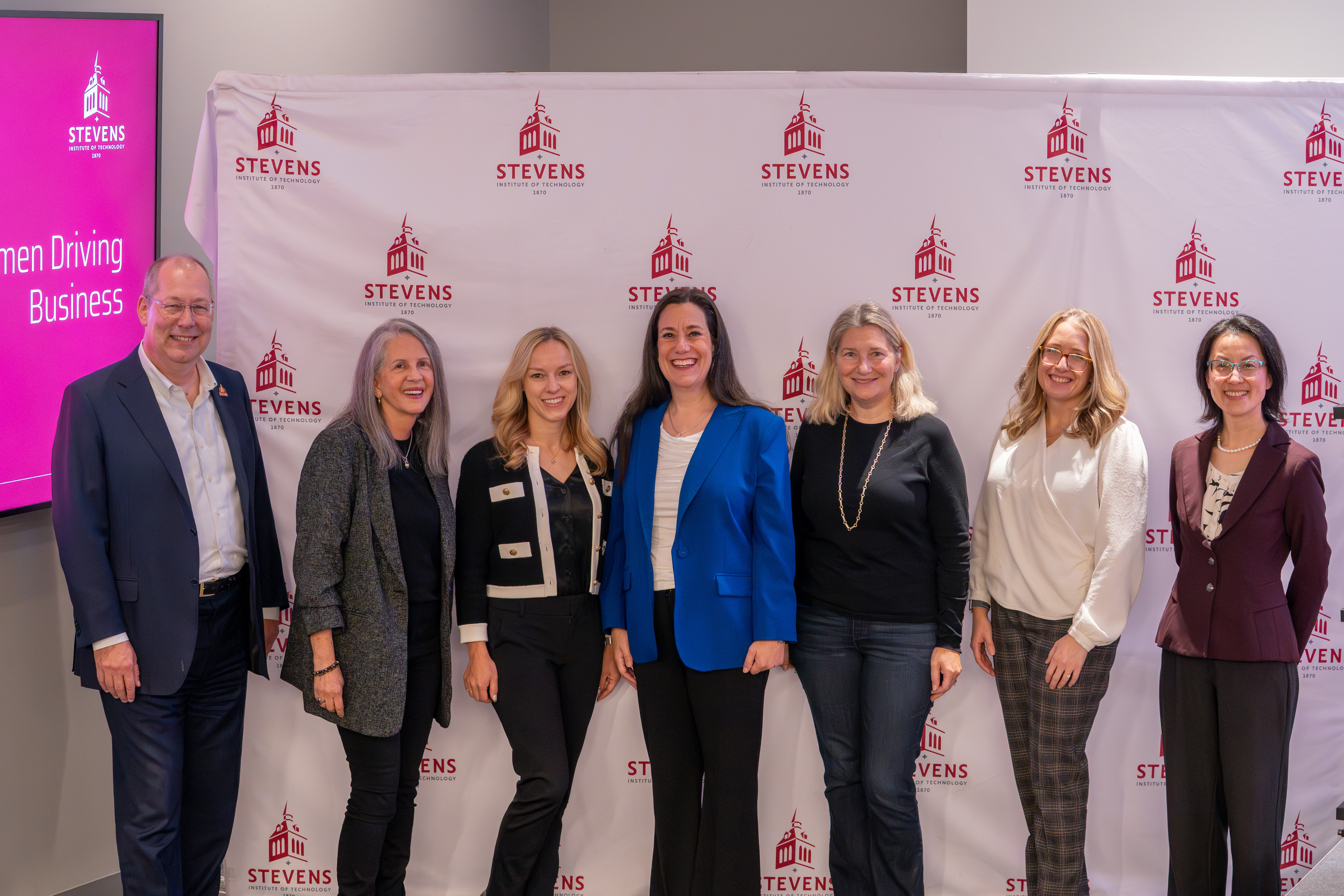 Seven business professionals, mostly women, pose together in front of a Stevens Institute of Technology backdrop. A pink screen displaying "Women Driving Business" is visible on the left. The group appears to be at a panel or networking event.