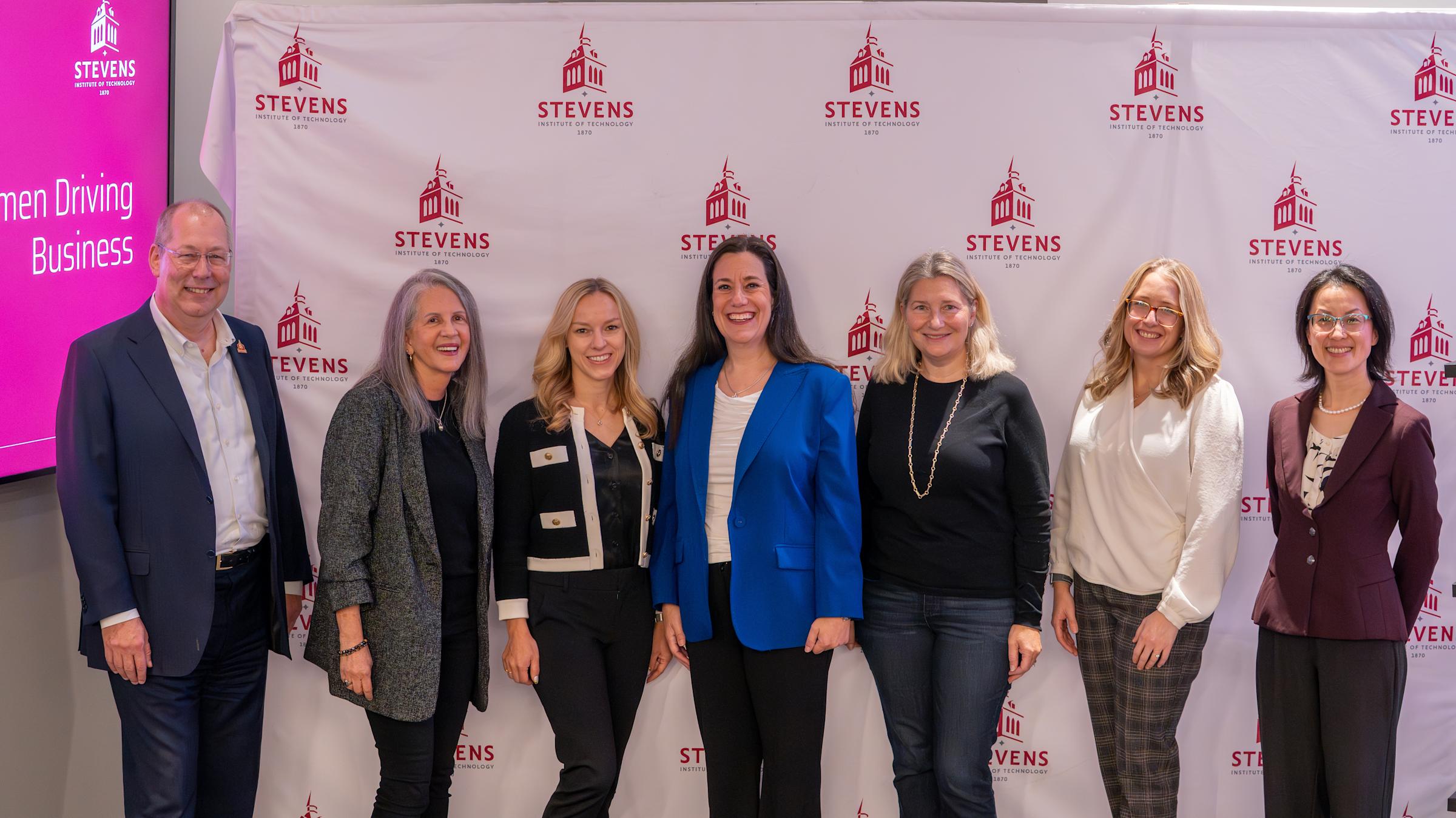 Seven business professionals, mostly women, pose together in front of a Stevens Institute of Technology backdrop. A pink screen displaying "Women Driving Business" is visible on the left. The group appears to be at a panel or networking event.