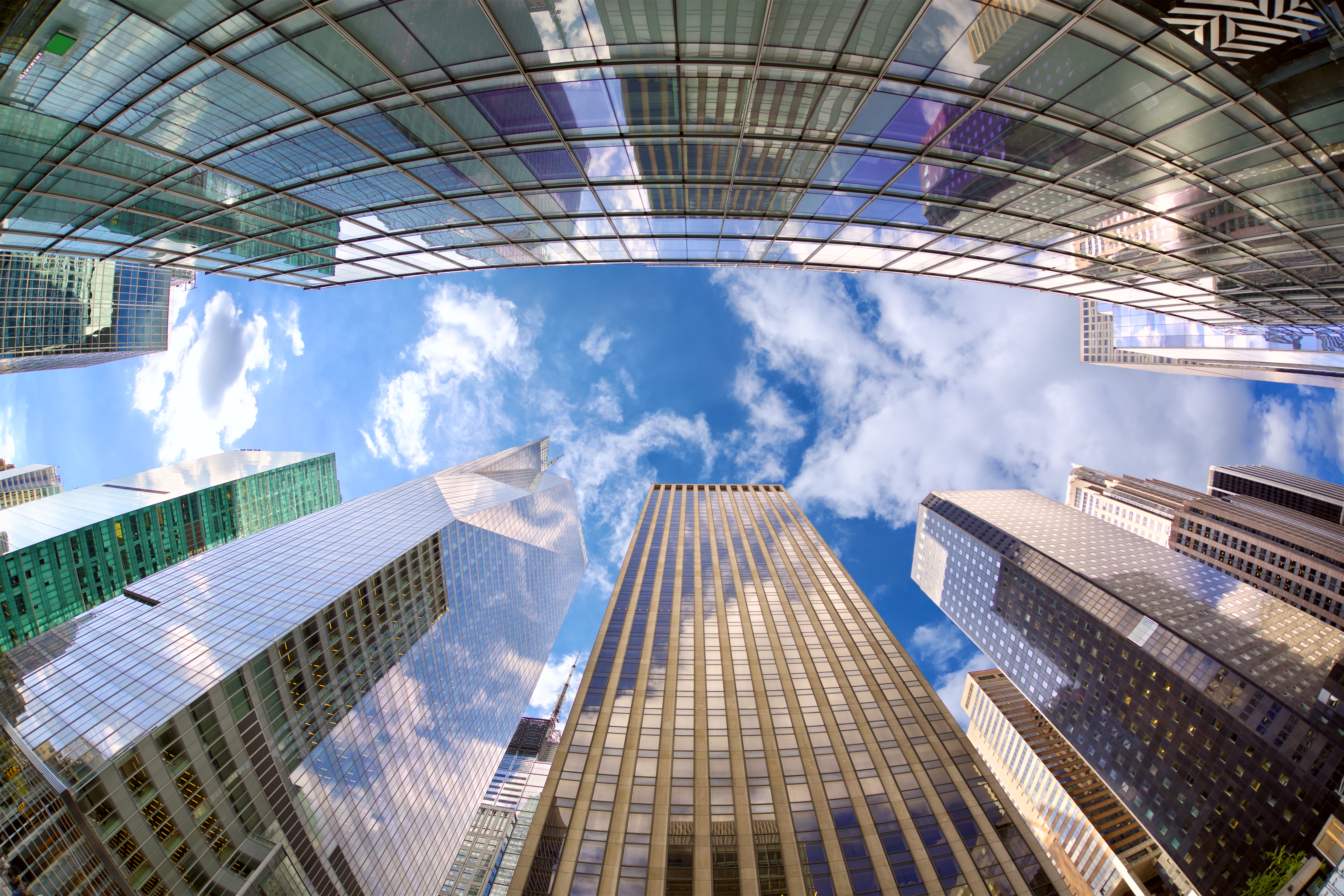A view of midtown Manhattan skyscrapers from street level looking straight up into a blue sky with white clouds.