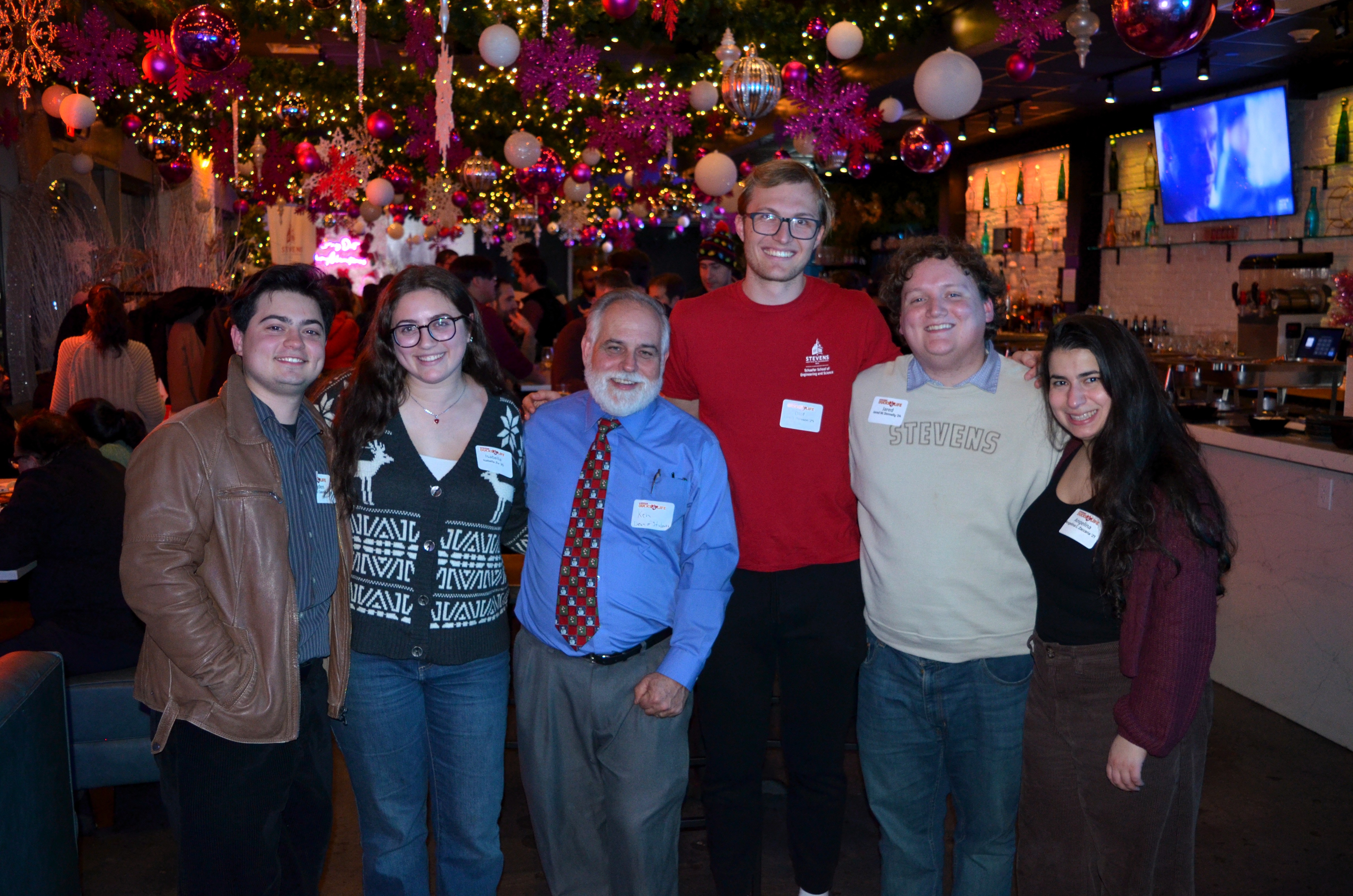 Six attendees pose together in a decorated restaurant at the SAA Holiday Gathering in 2025. Colorful ornaments, snowflakes and string lights hang overhead, while guests mingle in the background.