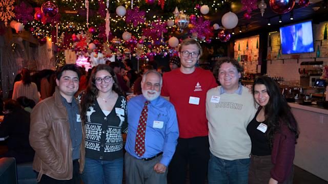 Six attendees pose together in a decorated restaurant at the SAA Holiday Gathering in 2025. Colorful ornaments, snowflakes and string lights hang overhead, while guests mingle in the background.