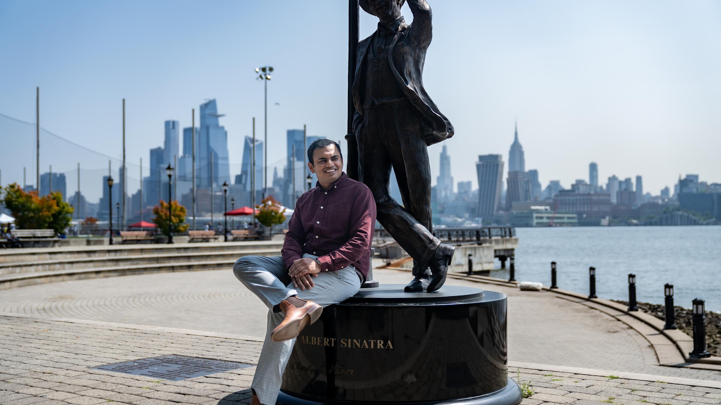 Mani sits on the Frank Sinatra statue in Hoboken with part of the New York City skyline behind him.