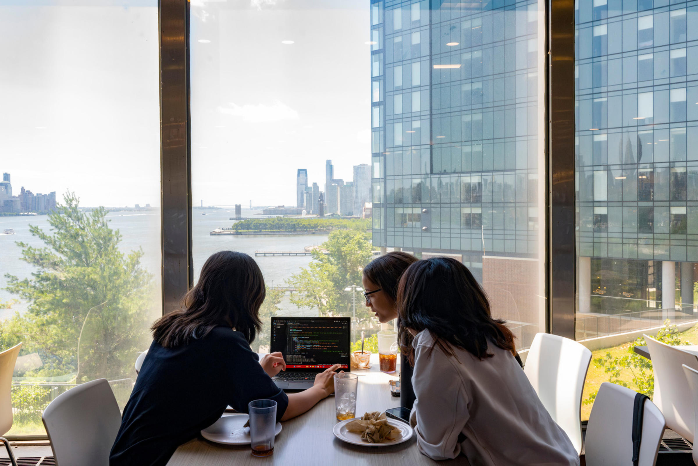 Three high school students look at a small computer monitor in front of a glass window with the Hudson River in the background.