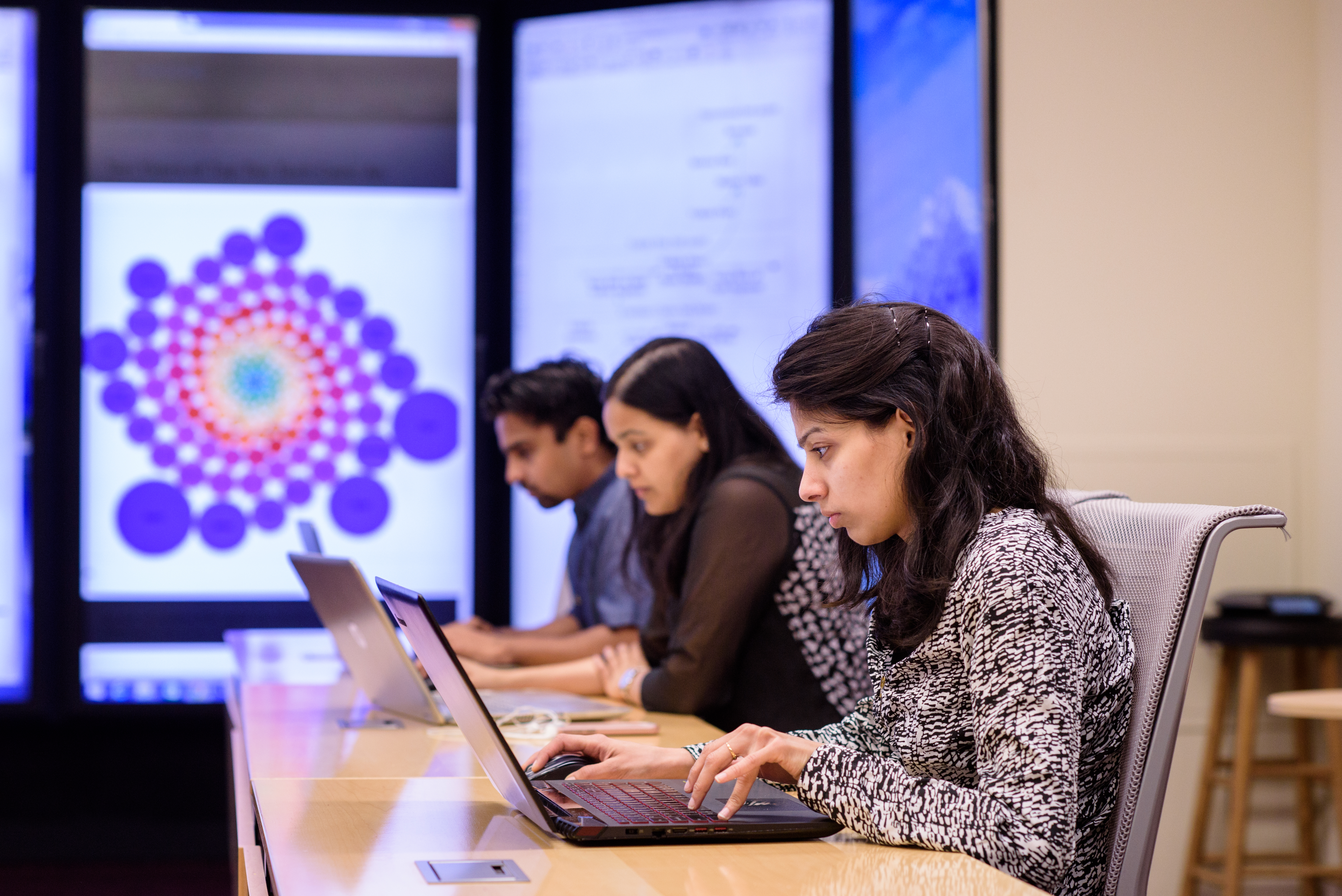A student working on her laptop with screen of visual data in background.