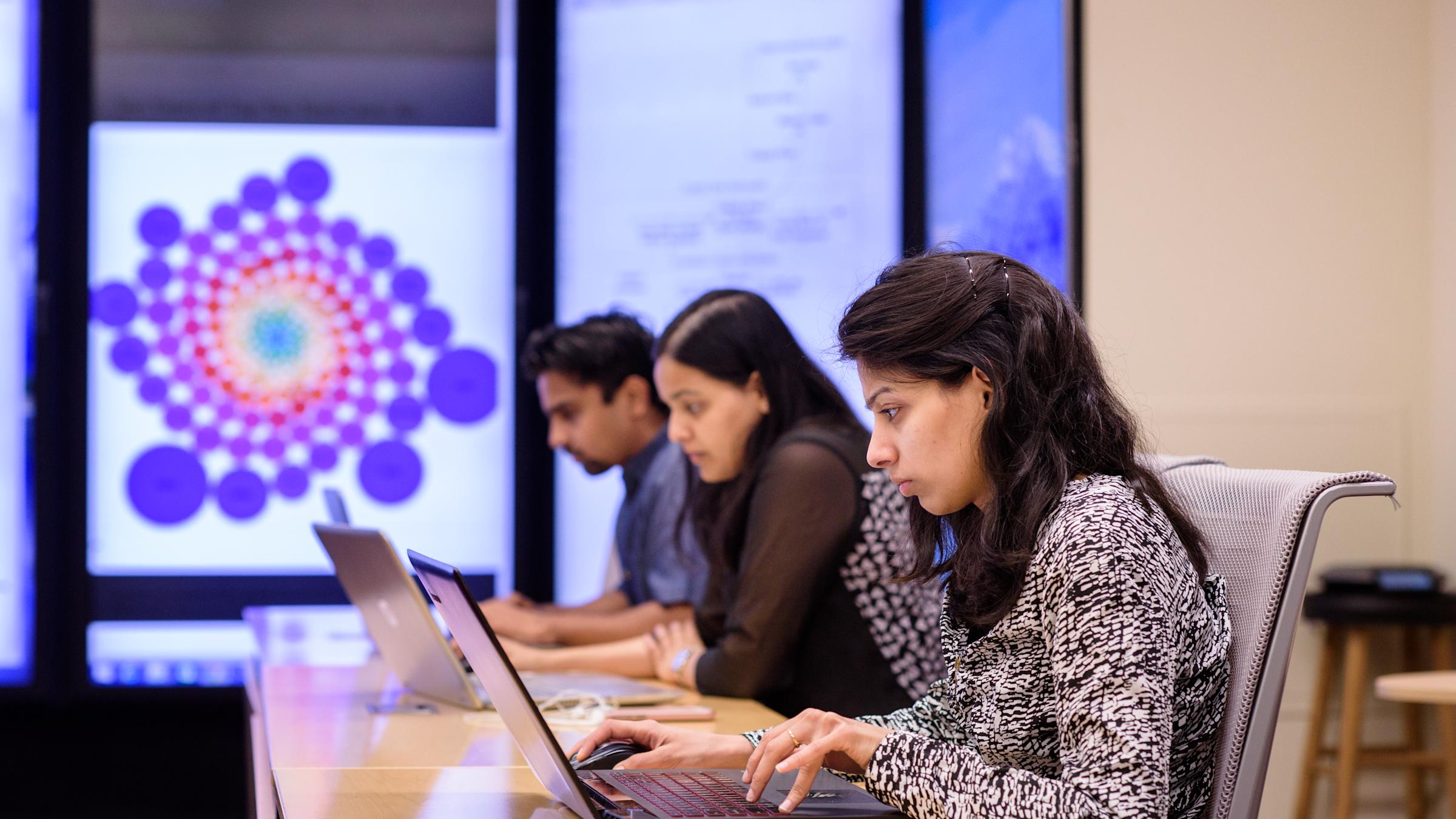 A student working on her laptop with screen of visual data in background.