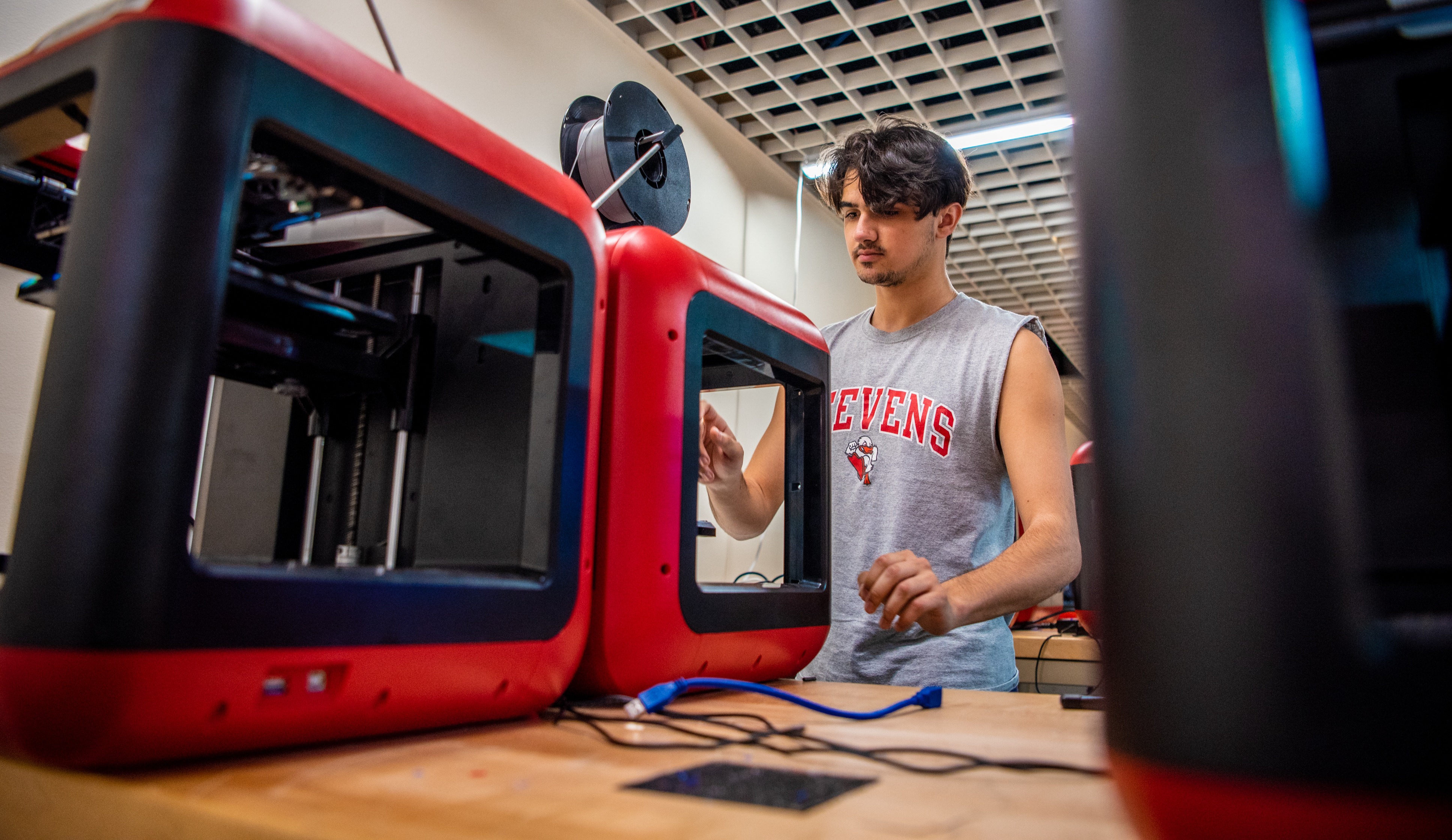 A male student in a Stevens muscle t-shirt works with equipment in a robotics lab.