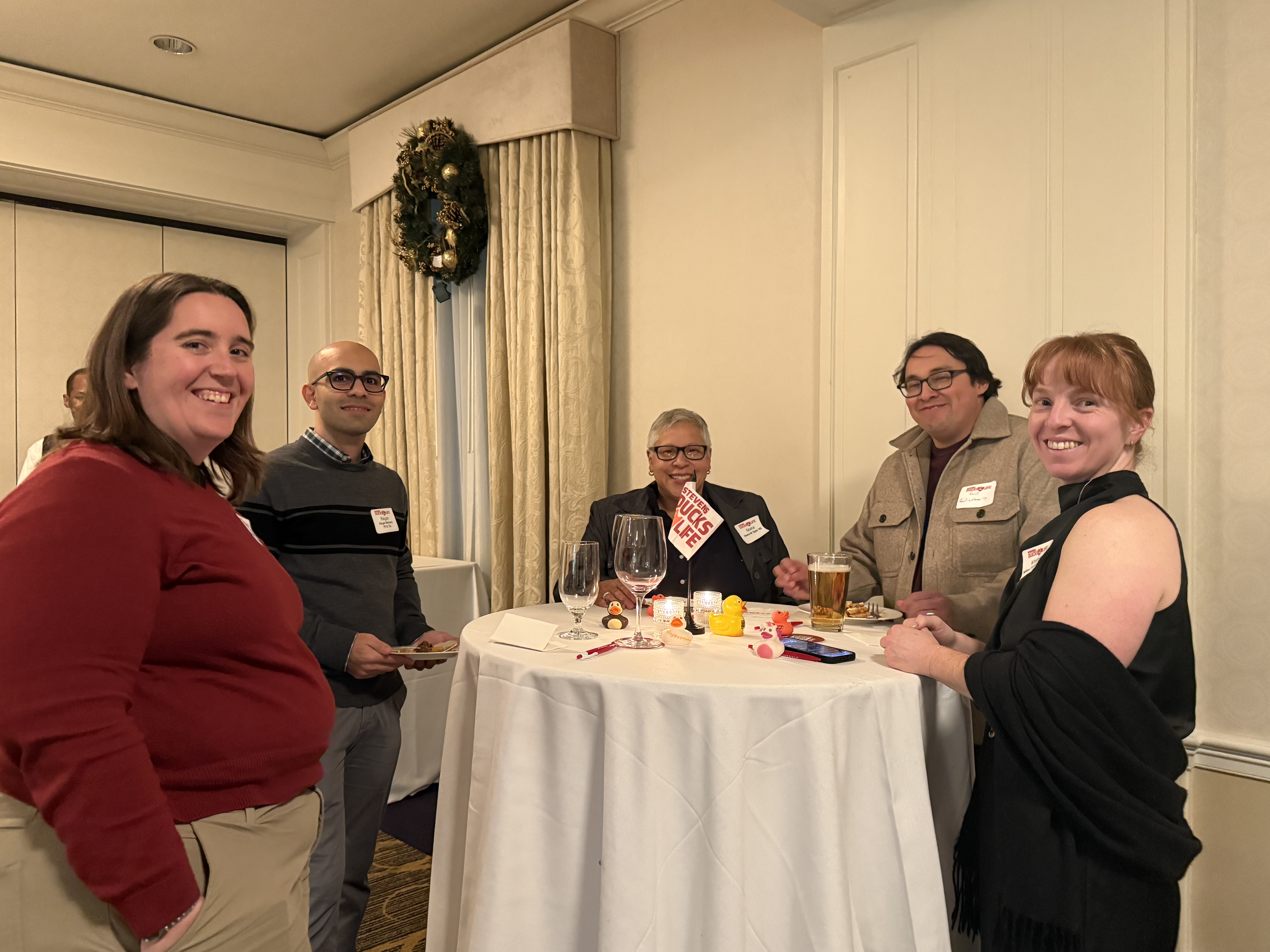Five attendees gather around a cocktail table at the Washington, D.C. Alumni Holiday Gathering 2025, enjoying conversation in a warmly lit event space decorated with seasonal holiday wreaths and table accents.
