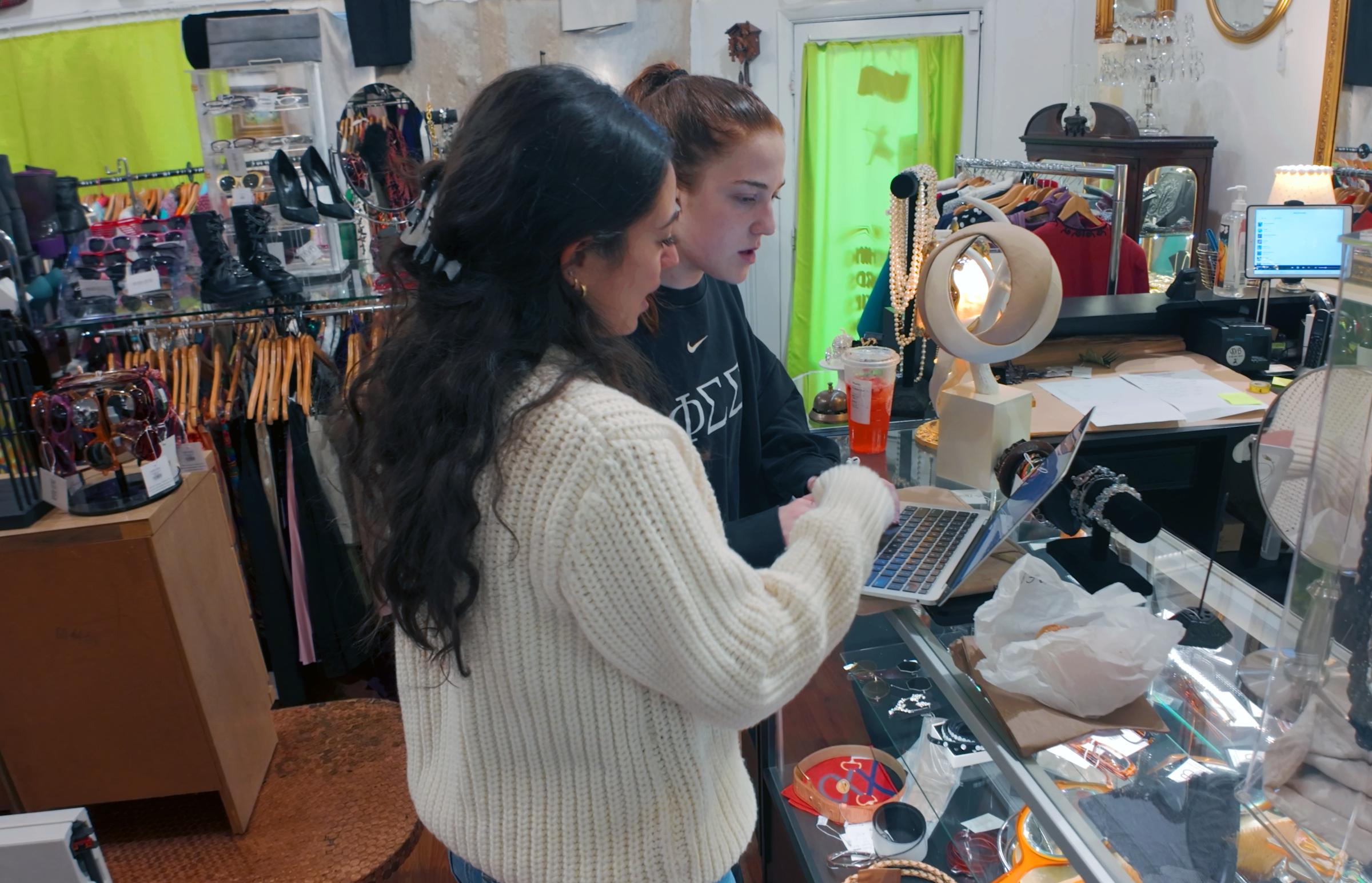 Two students work at a laptop at the counter of a vintage clothing store. 