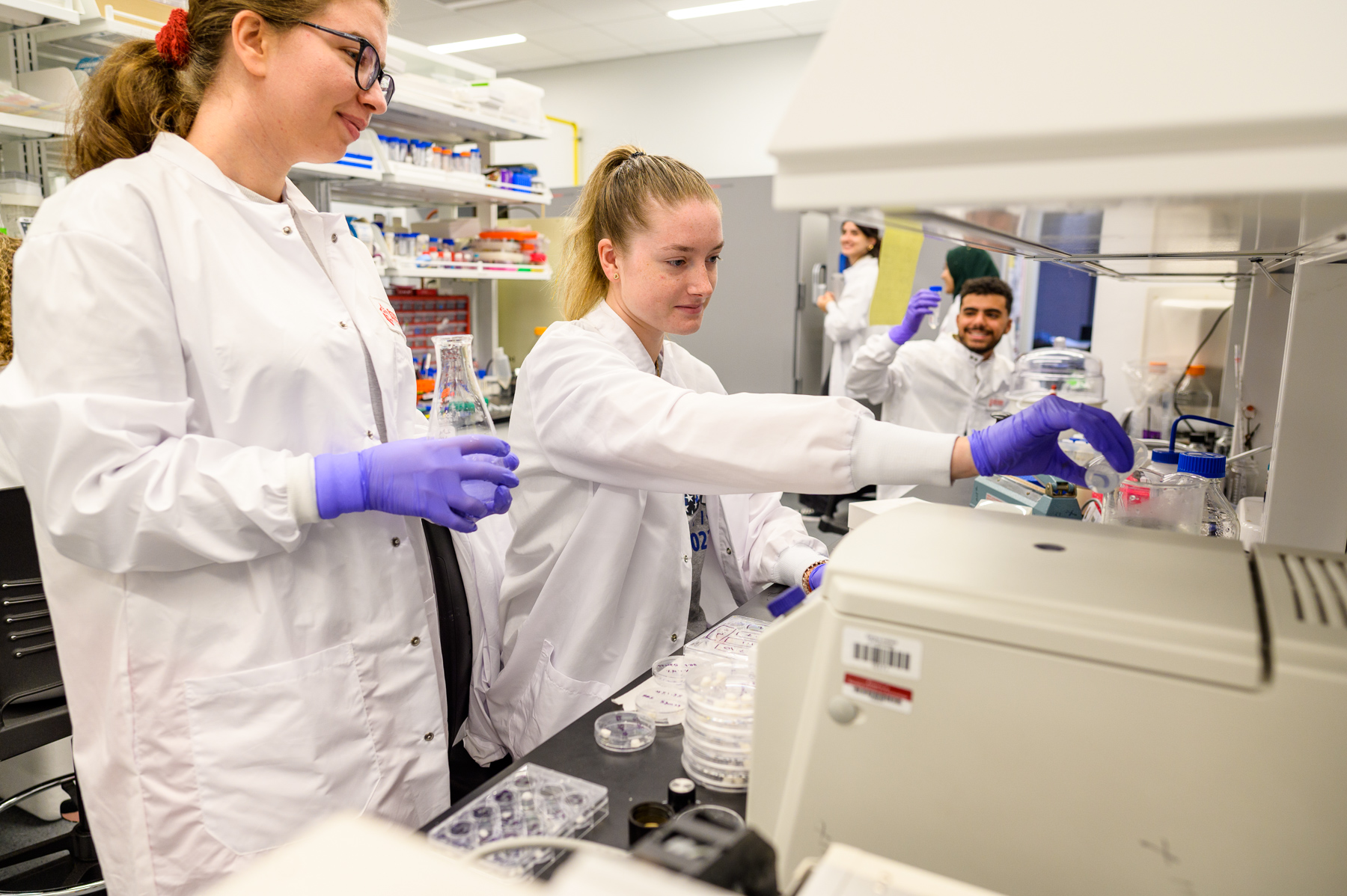 Two women students dressed in lab coats work in a lab.