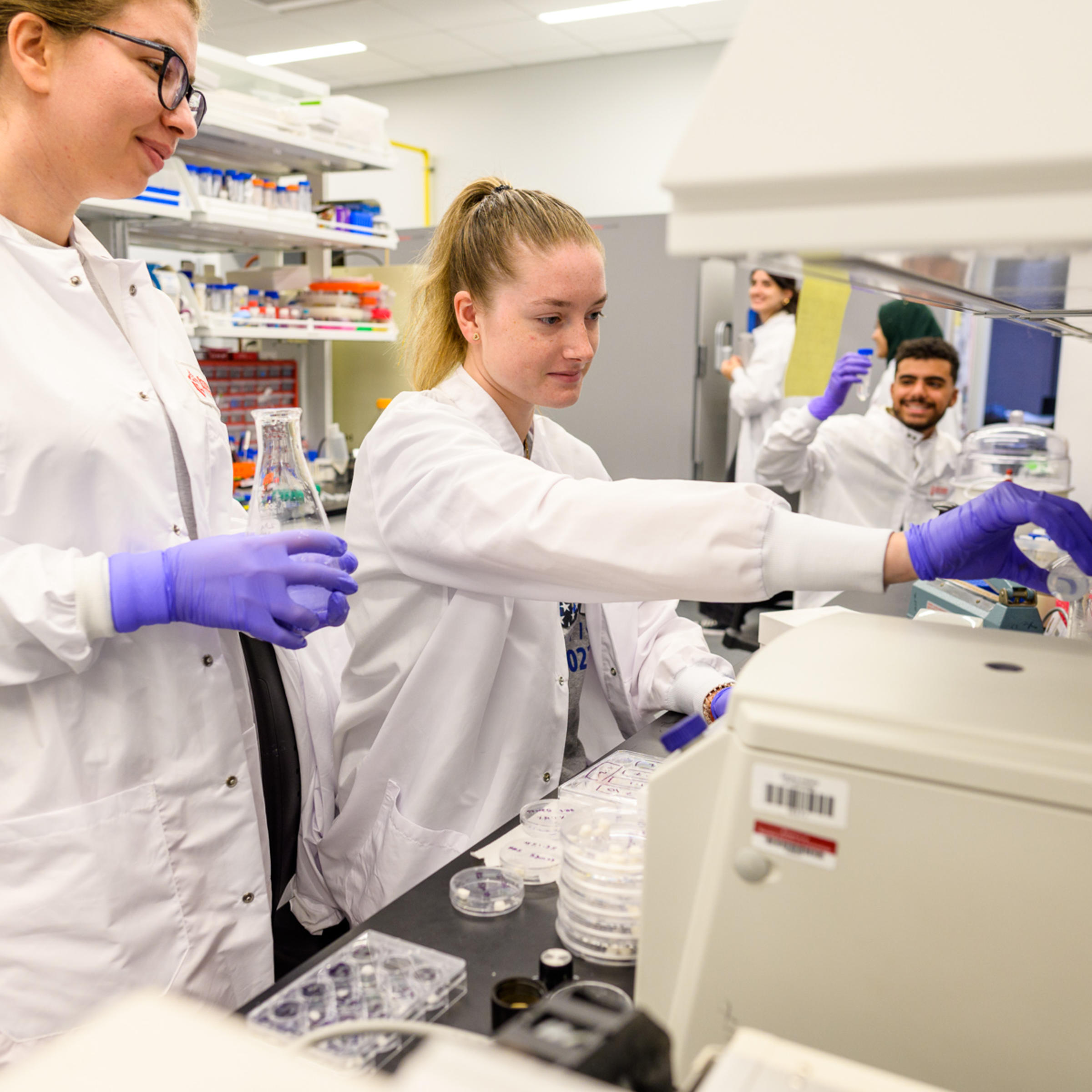 Two women students dressed in lab coats work in a lab.