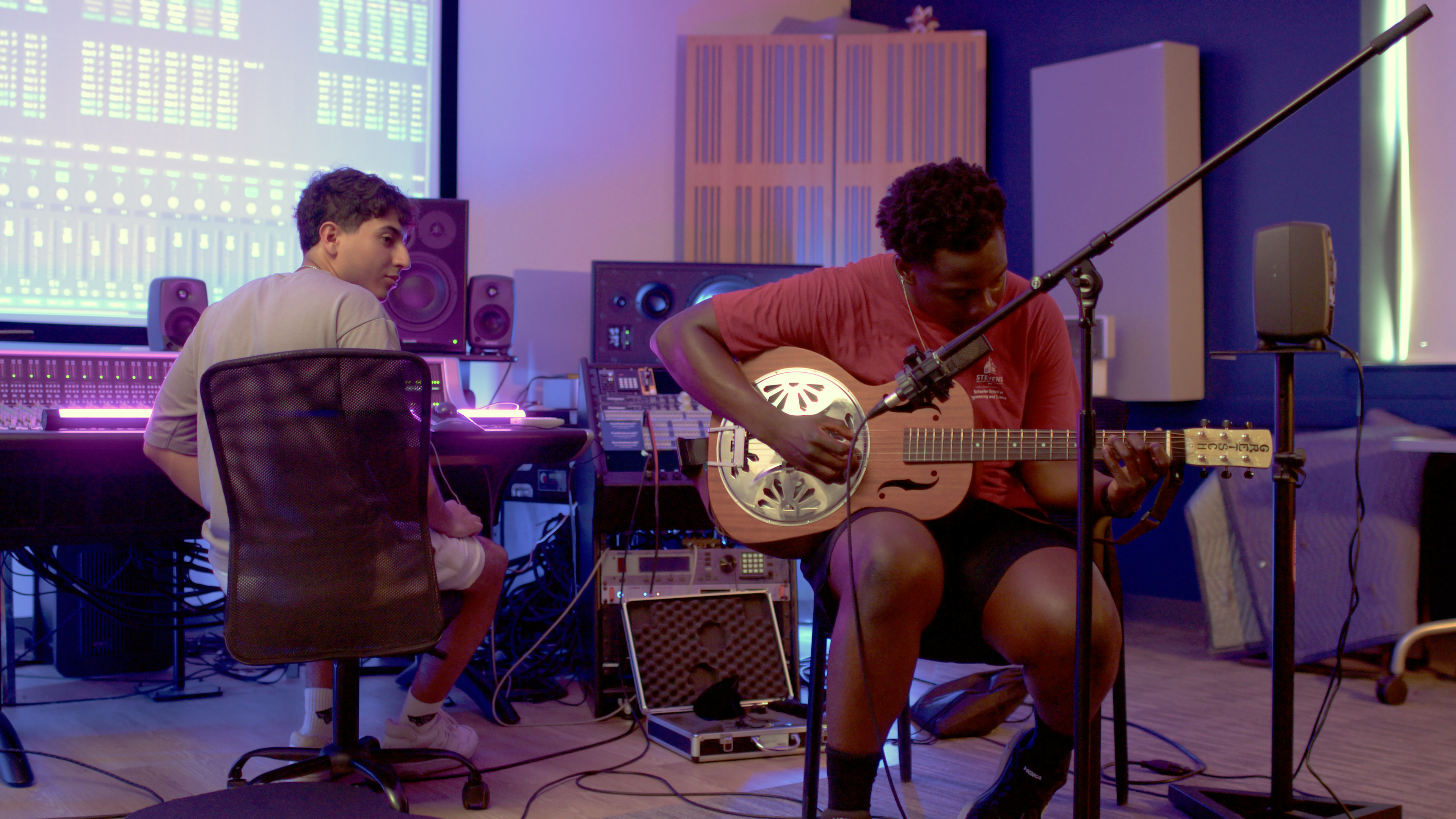 Two students in a sound lab making music. One is playing guitar, the other is at a mixing board watching the guitarist.