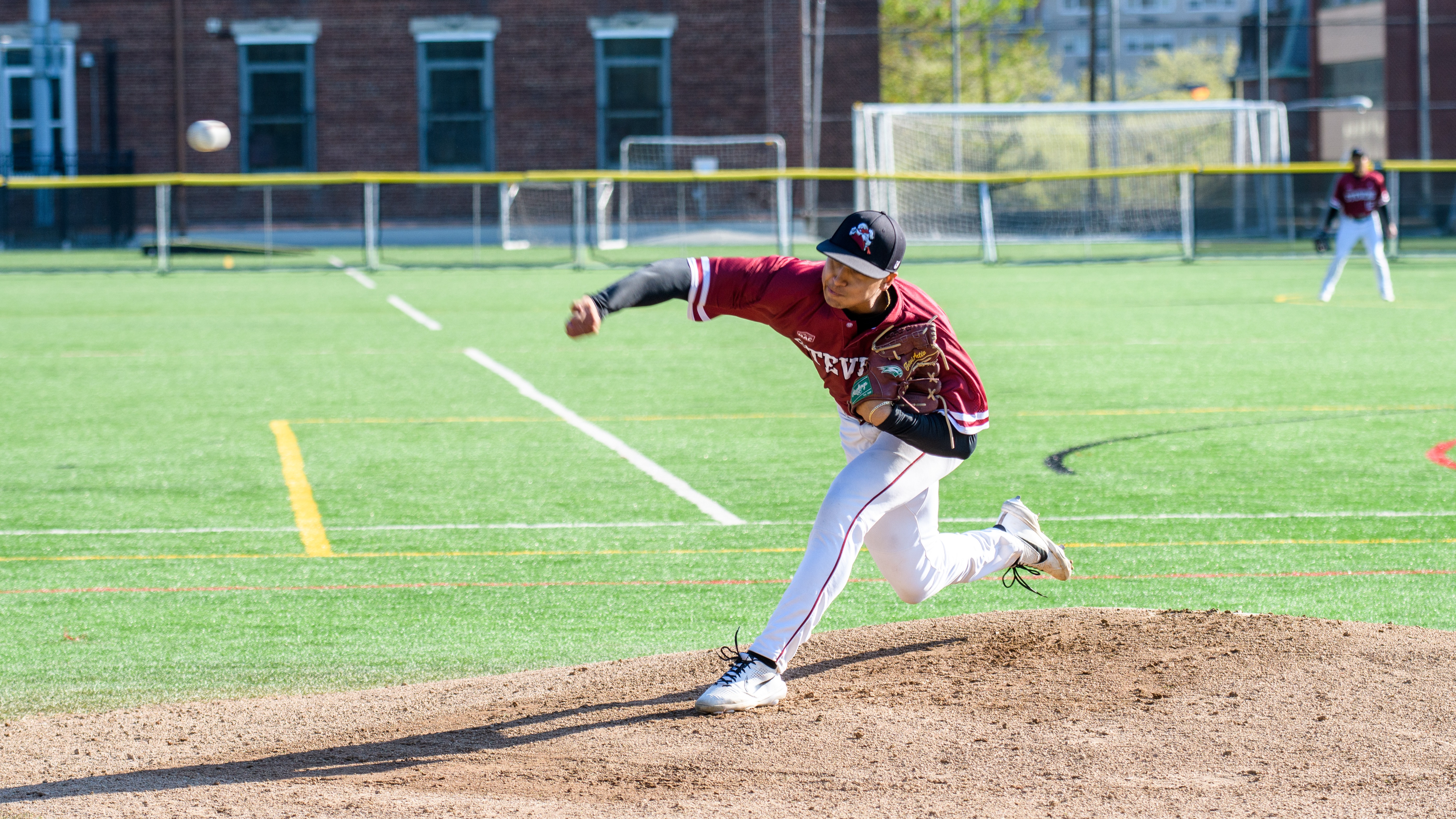 A right-handed baseball pitcher for Stevens releases a pitch off the mound with the baseball out of his hand and in frame on its way to home plate.