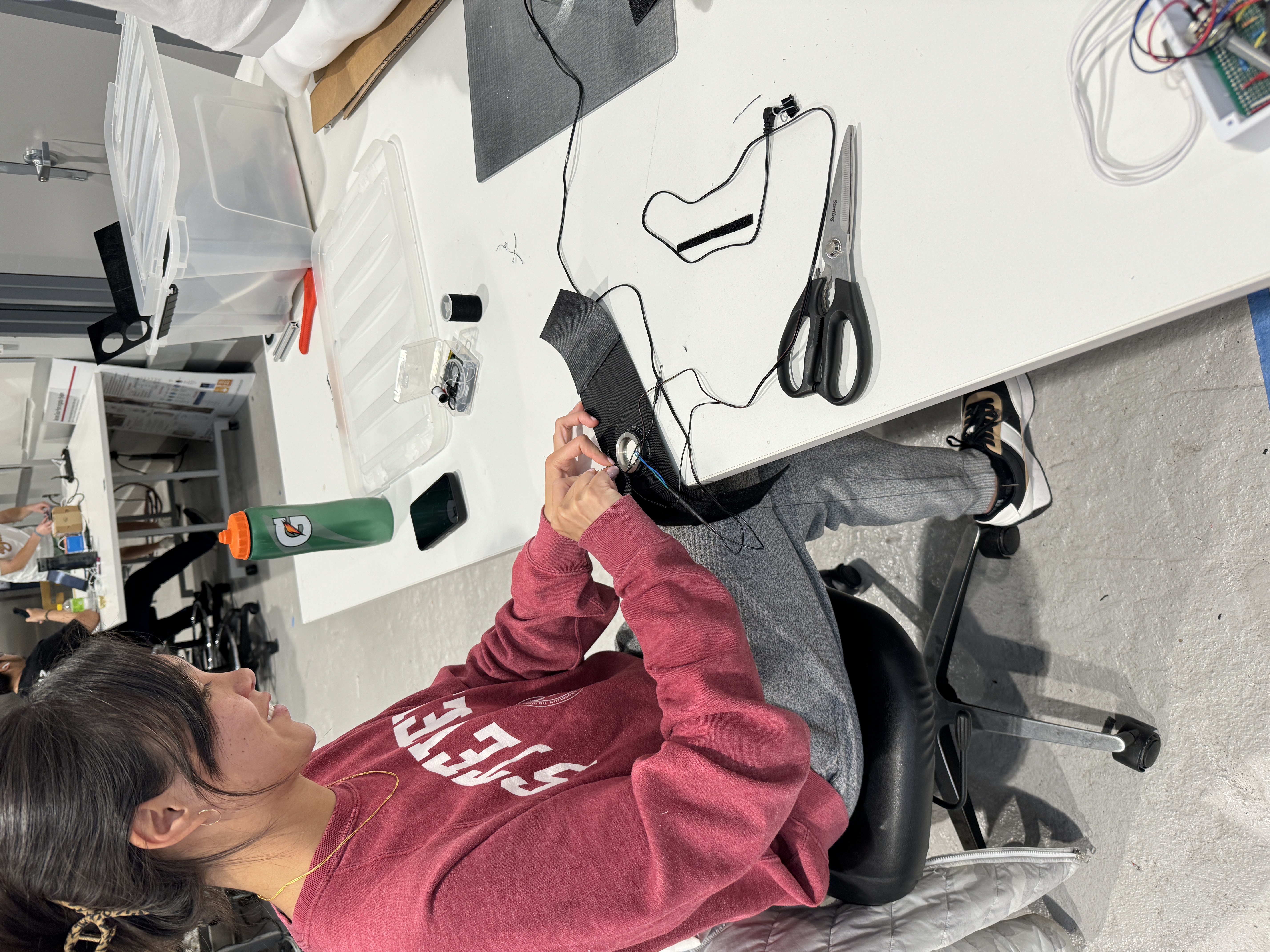 A female student in a red Stevens sweatshirt sits at a table in a lab with a pair of scissors next to her. In her hands she is working on assembling the Medflex device.