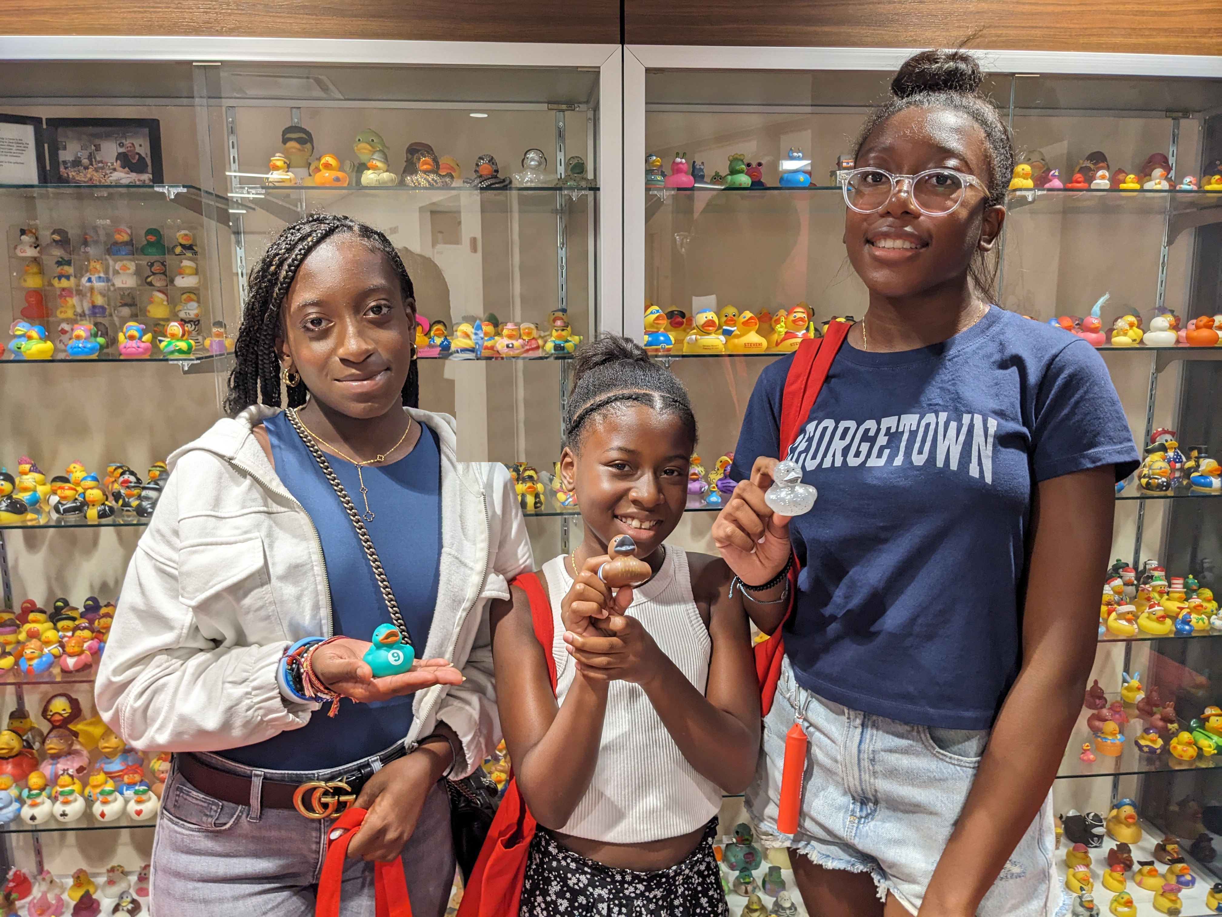 Three young women holding rubber ducks in front of a glass display case containing more ducks.
