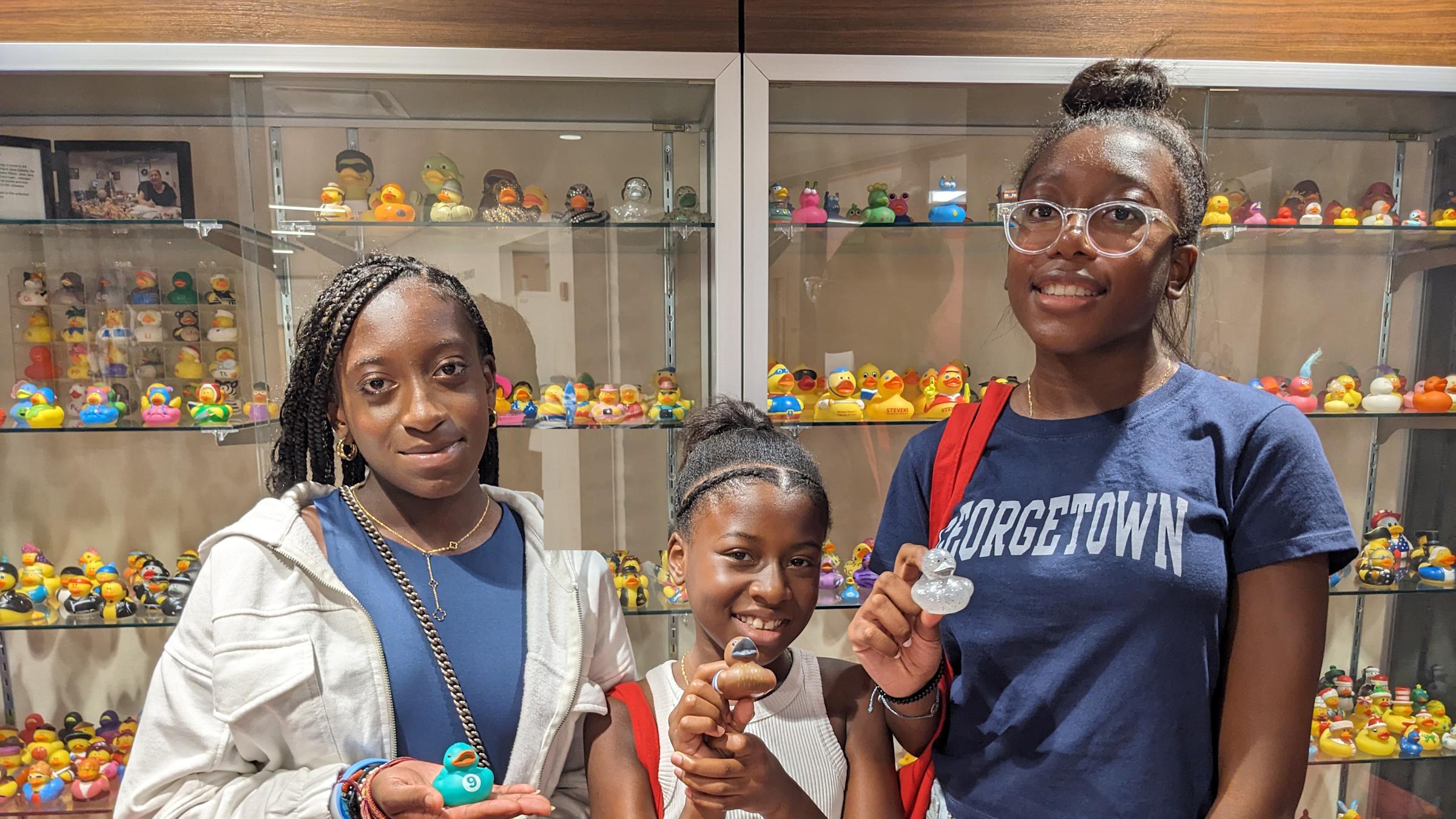 Three young women holding rubber ducks in front of a glass display case containing more ducks.