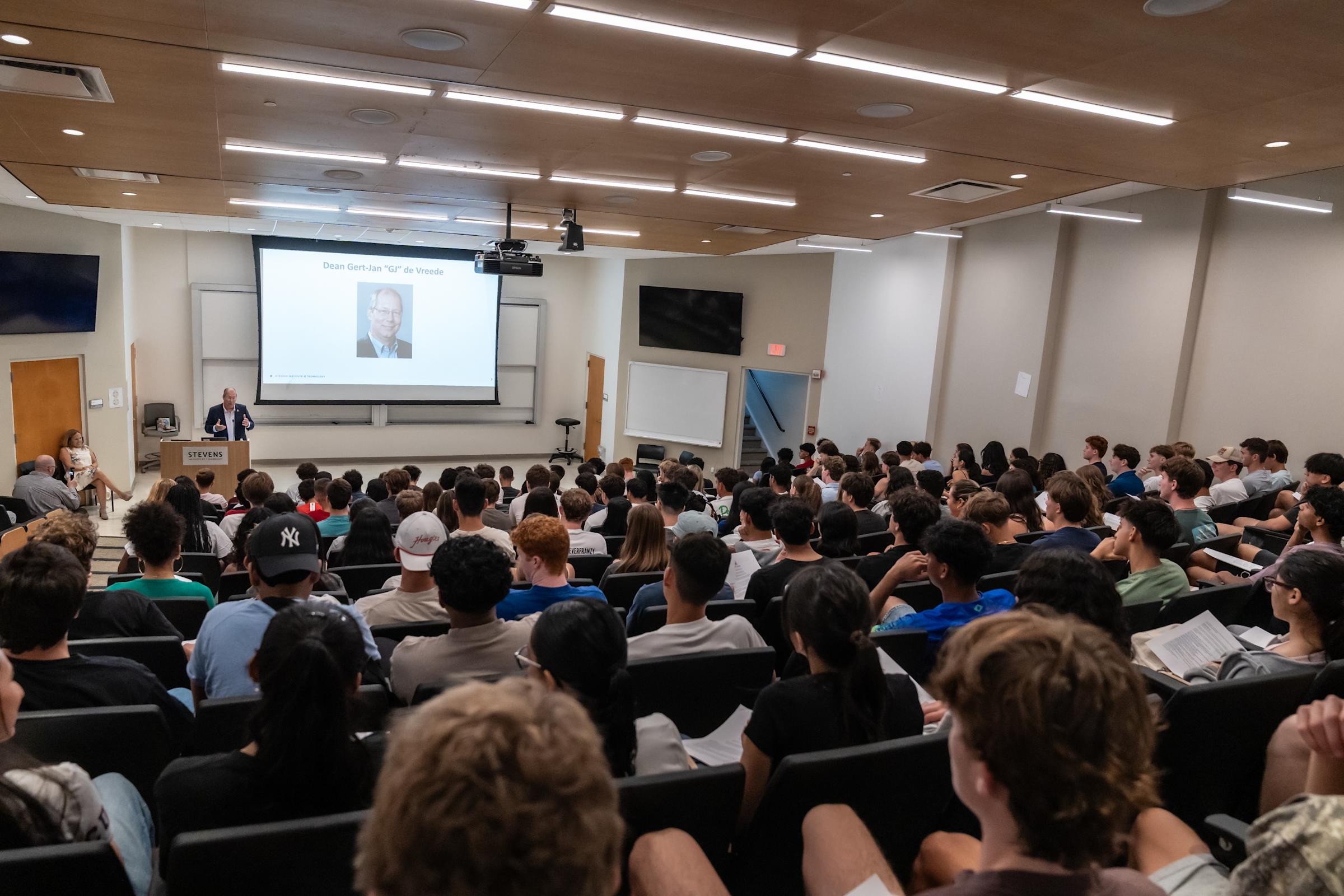 Packed university lecture hall with students in tiered seating watching professor present at podium, large screen displaying speaker's photo.