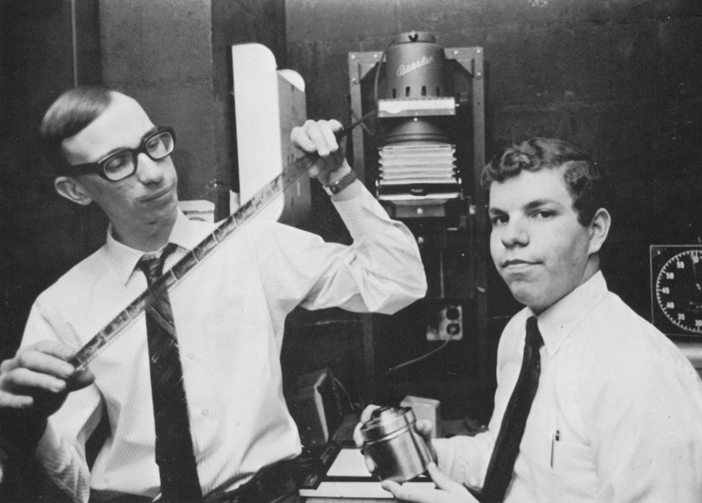 Aged black and white photo of Donald Silawsky and friend looking at photo negatives in darkroom.