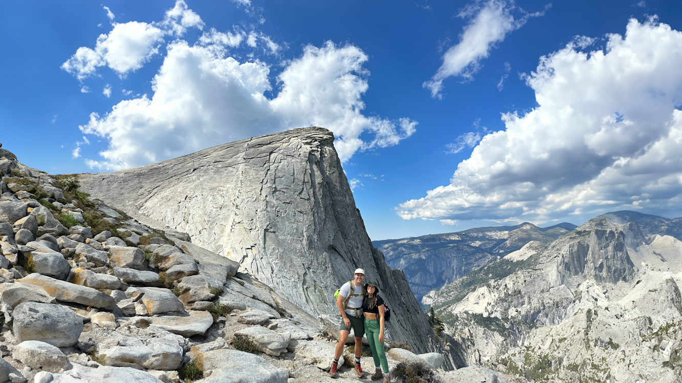 A man and a woman stand at the summit of a mountain, surrounded by rocks, a mountain range, with a blue sky and clouds above them.