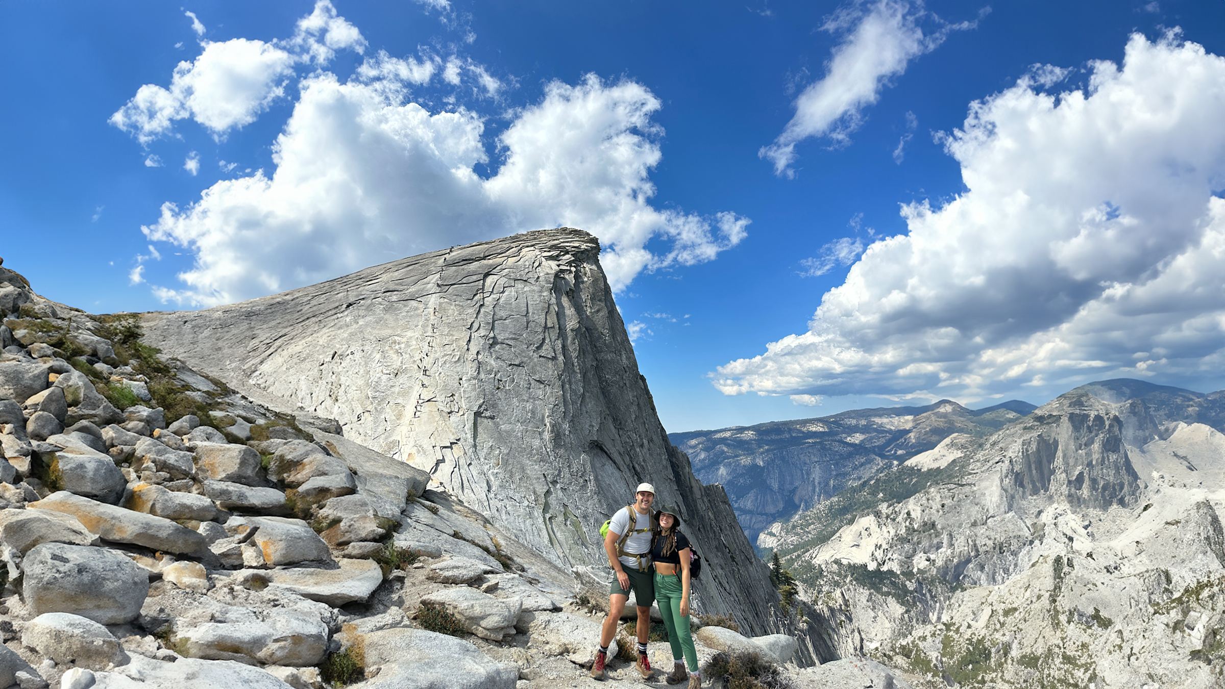 A man and a woman stand at the summit of a mountain, surrounded by rocks, a mountain range, with a blue sky and clouds above them.