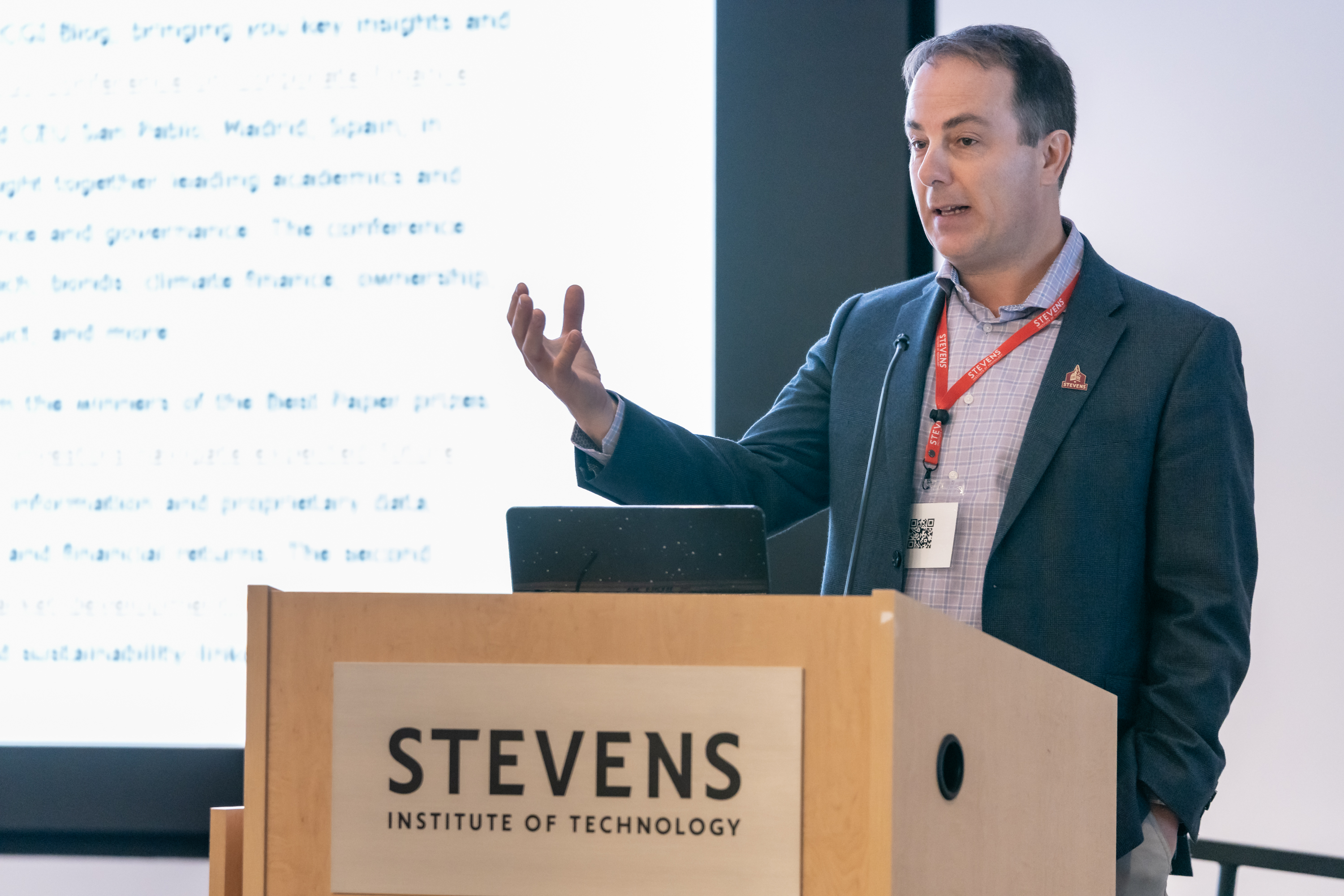 A man in a dark blazer and checkered shirt with a red lanyard presents at a wooden podium bearing the Stevens Institute of Technology logo. He gestures while speaking, with a laptop and projected presentation visible behind him.