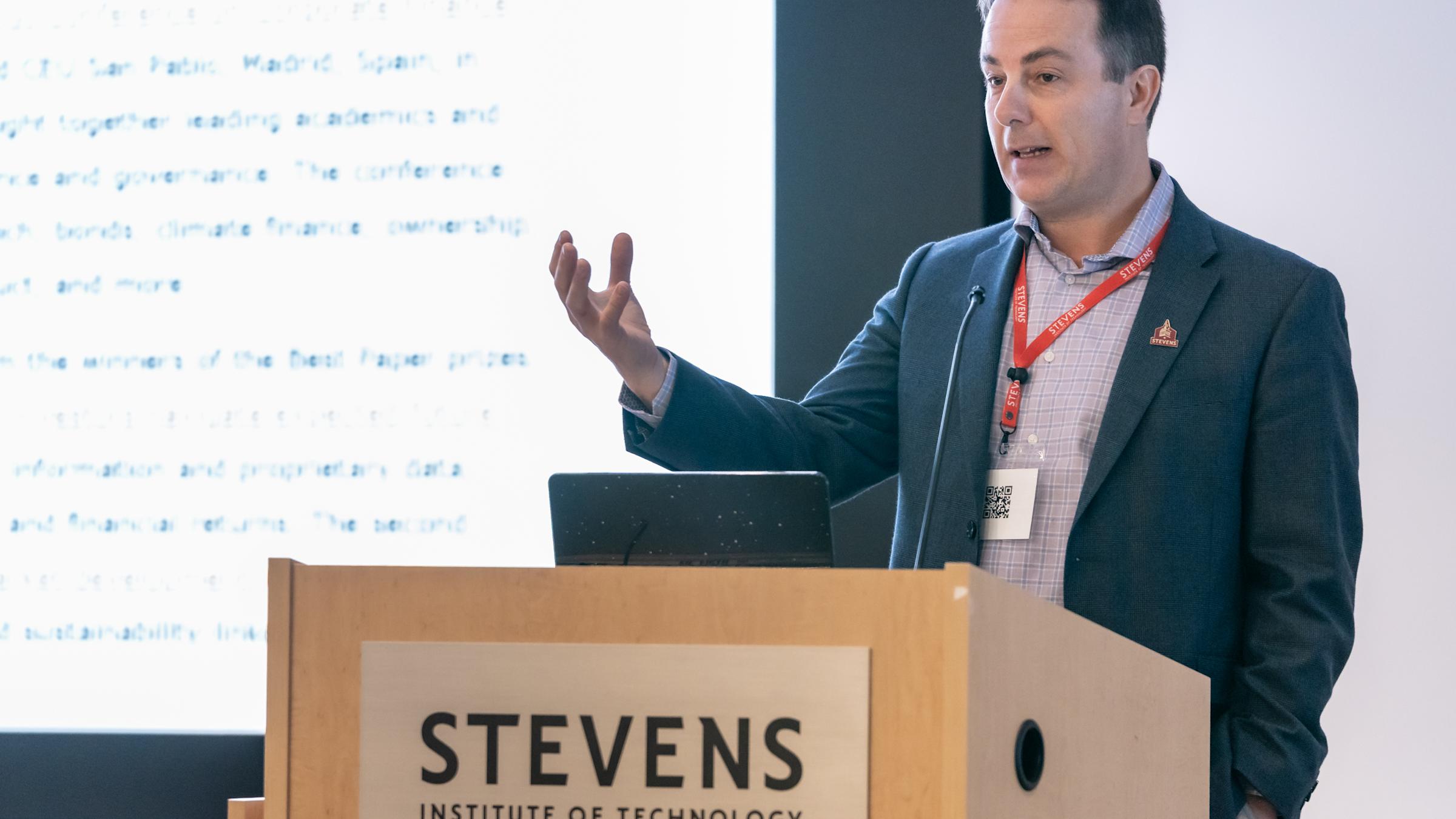 A man in a dark blazer and checkered shirt with a red lanyard presents at a wooden podium bearing the Stevens Institute of Technology logo. He gestures while speaking, with a laptop and projected presentation visible behind him.