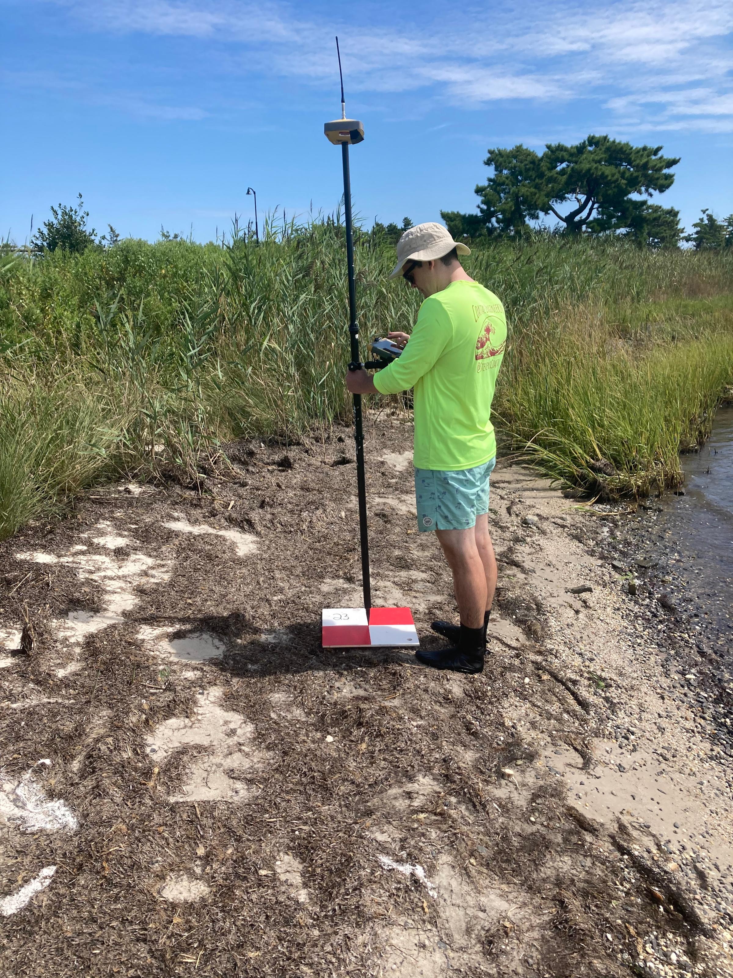 A man in a green shirt surveying elevation with a pole near a marshy area.