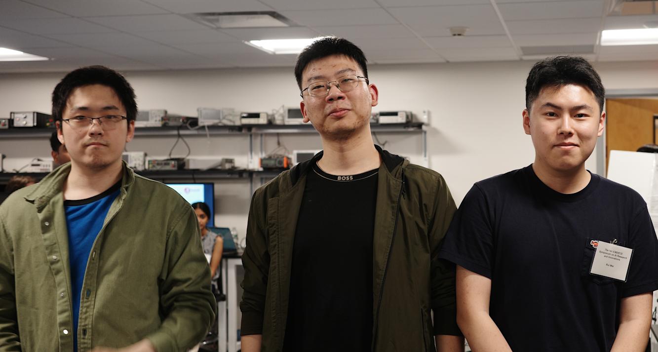 Three students stand next to each other in a lab looking at the camera. 