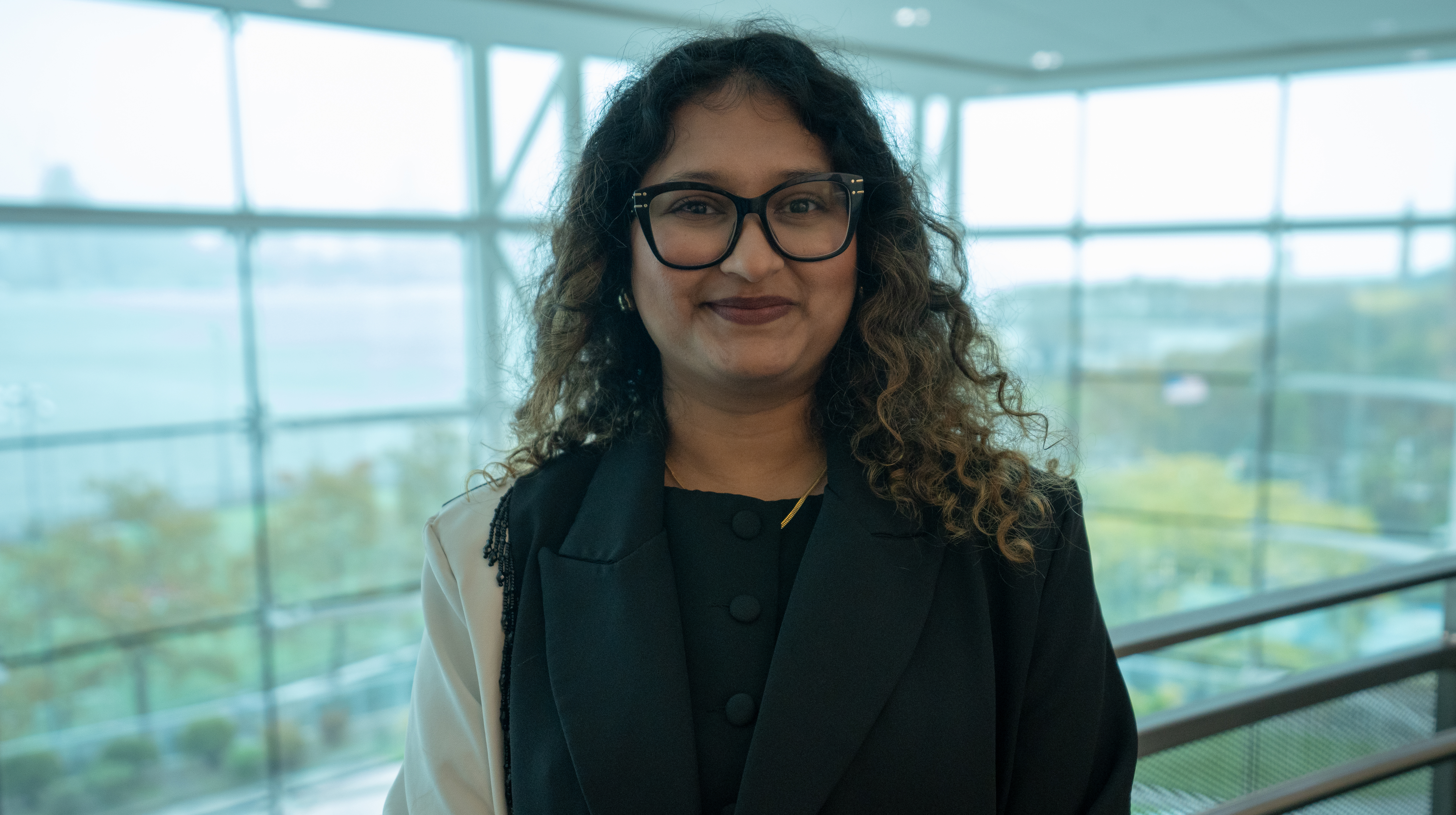A woman with curly dark hair and black-framed glasses wearing a dark blazer stands in a modern building interior with floor-to-ceiling windows overlooking a green campus area. She holds what appears to be a folder or portfolio and smiles at the camera.