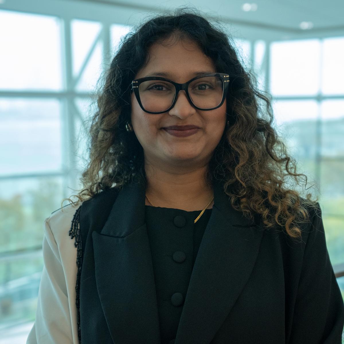 A woman with curly dark hair and black-framed glasses wearing a dark blazer stands in a modern building interior with floor-to-ceiling windows overlooking a green campus area. She holds what appears to be a folder or portfolio and smiles at the camera.