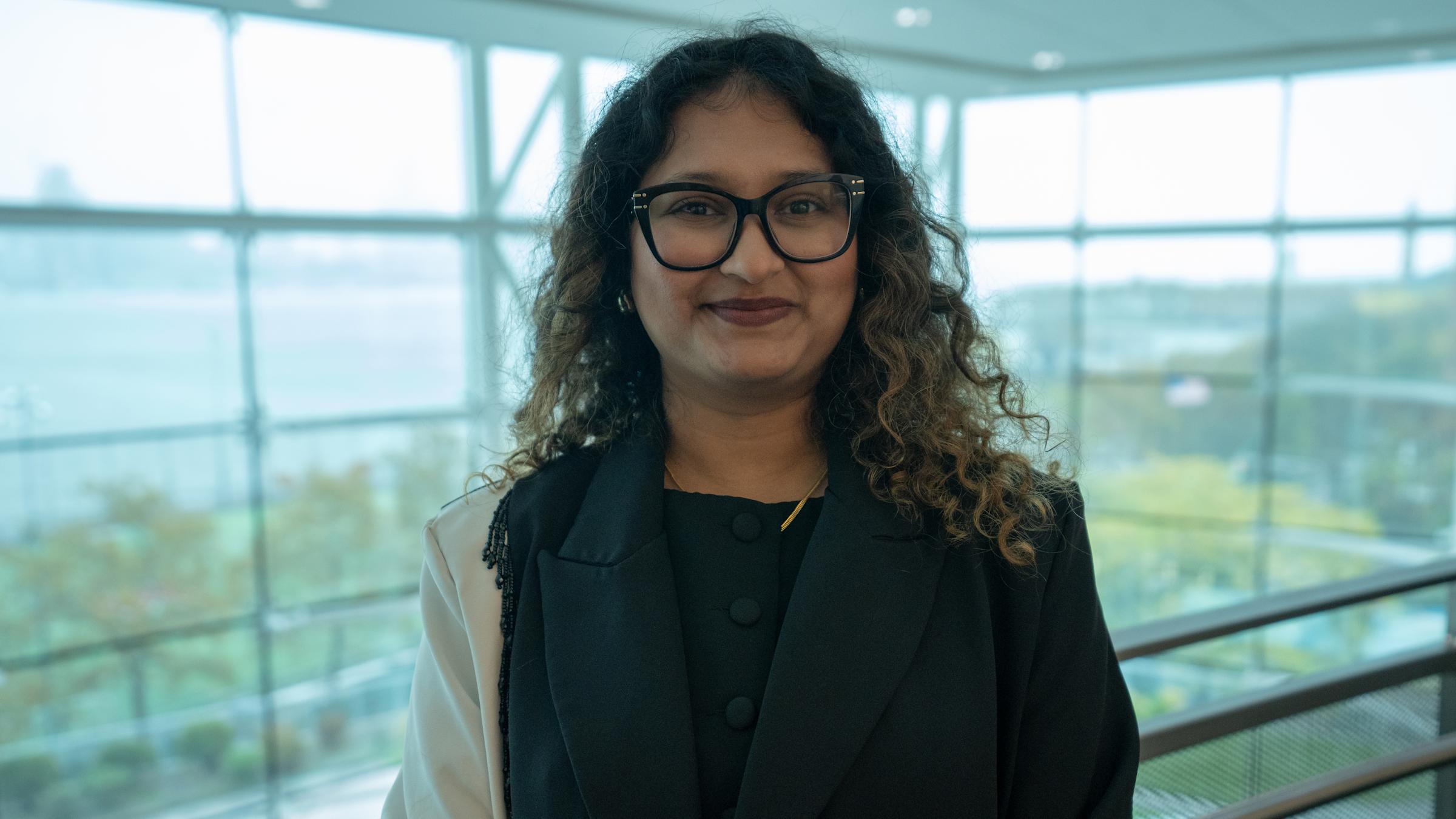 A woman with curly dark hair and black-framed glasses wearing a dark blazer stands in a modern building interior with floor-to-ceiling windows overlooking a green campus area. She holds what appears to be a folder or portfolio and smiles at the camera.