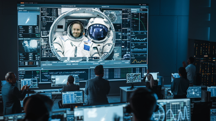 An image of a big screen in a busy control room with an astronaut featured on the screen and lots of graphs on the screen.