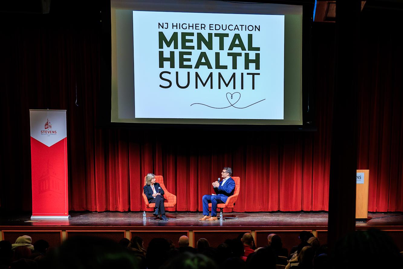 A man and woman sit on stage speaking in front of a projection screen.