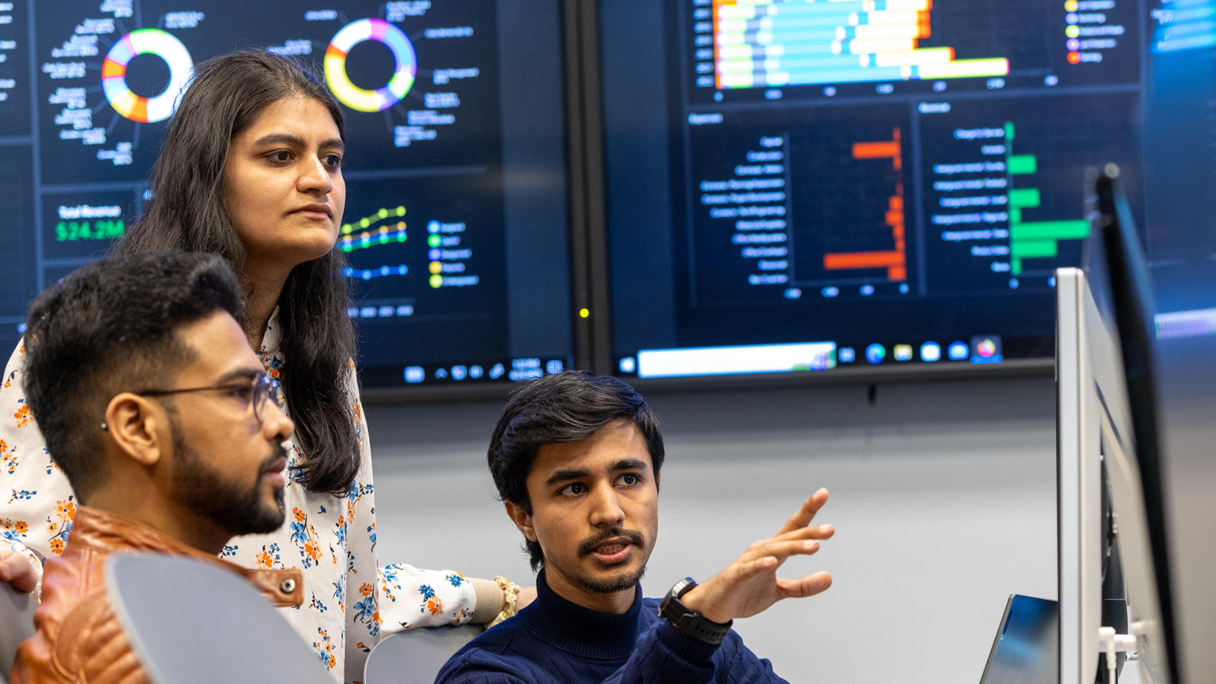 Three students view content on a monitor with data dashboards on screens behind them.