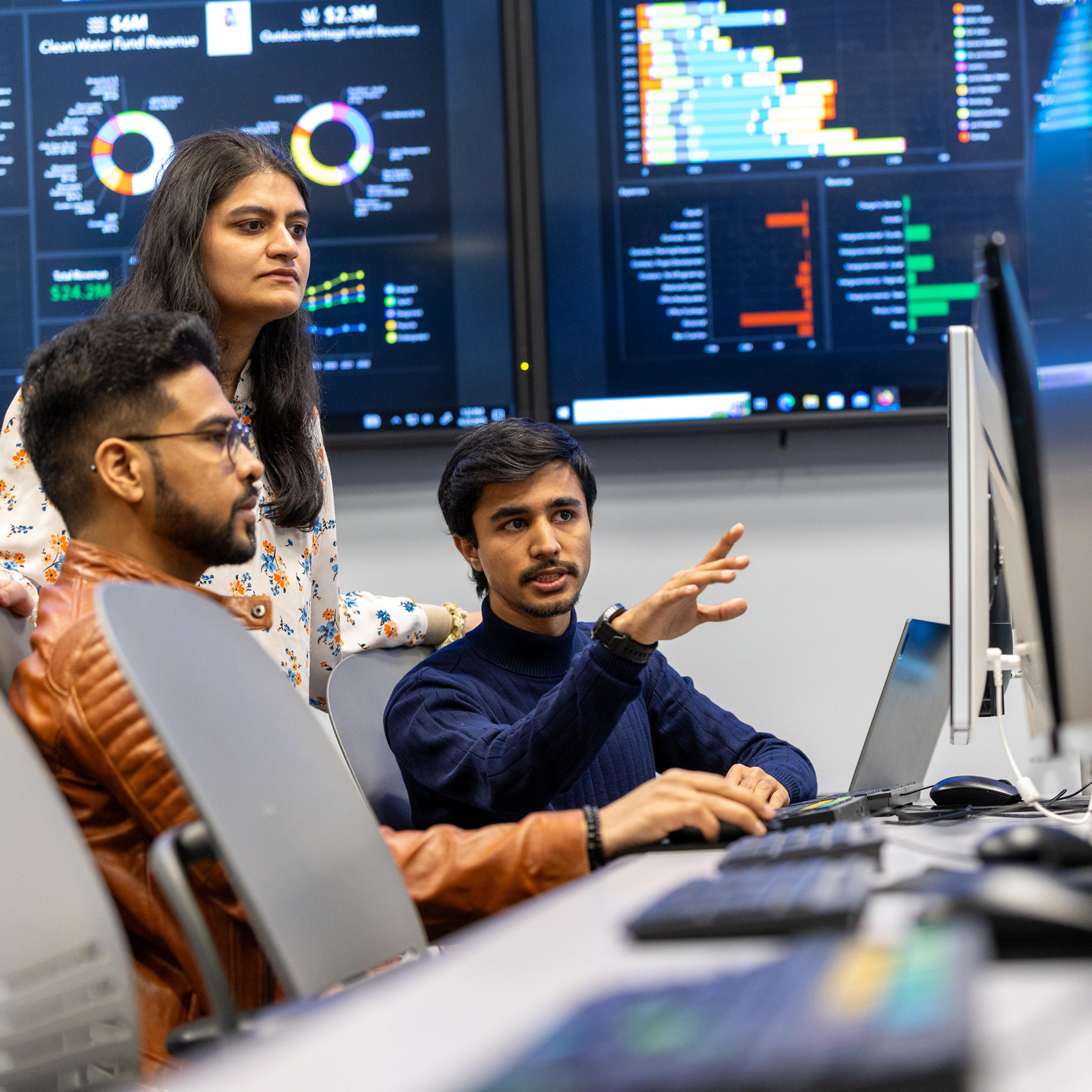 Three students view content on a monitor with data dashboards on screens behind them.